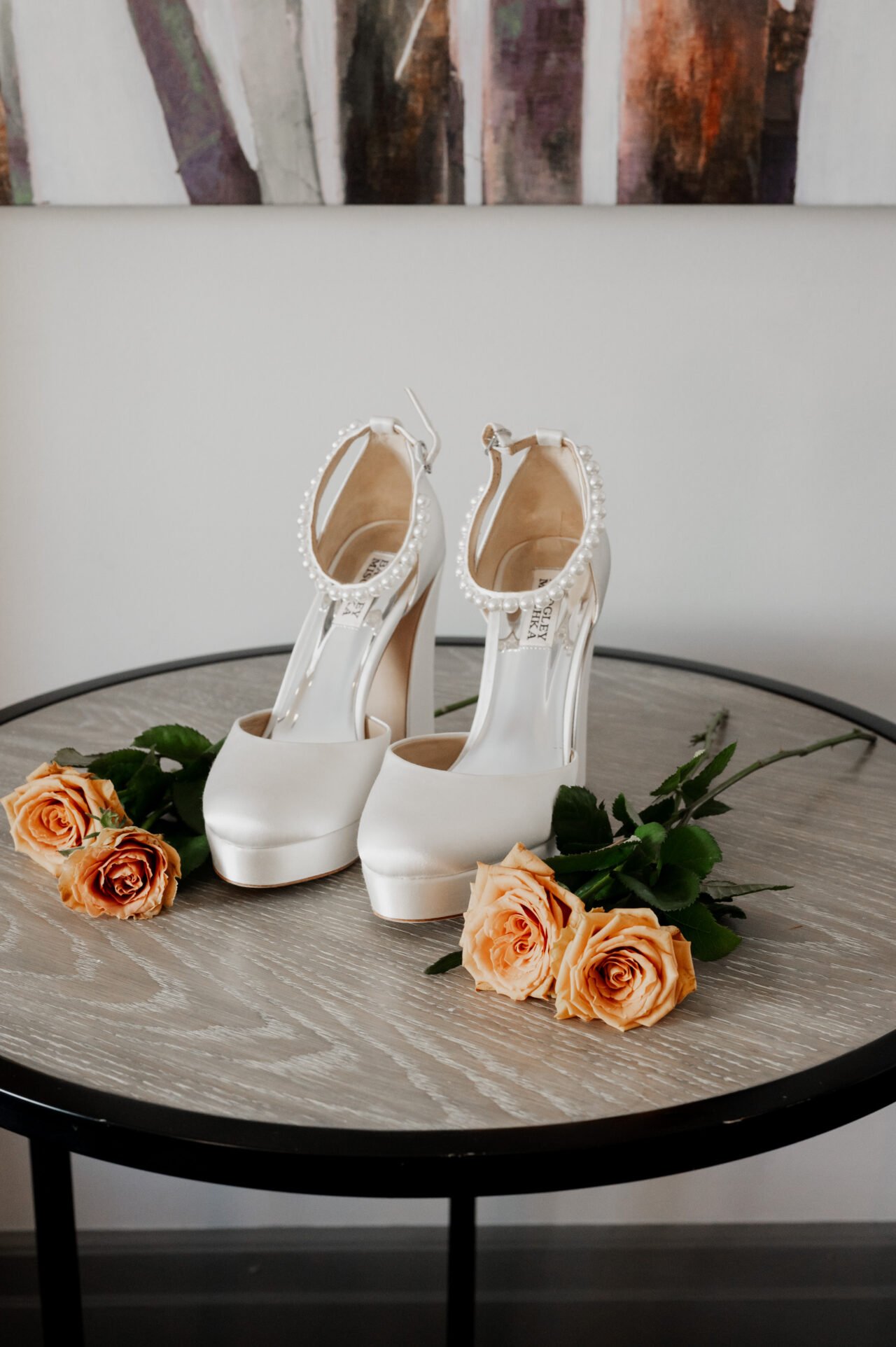 White high heels with embellishments sit elegantly on a round table, surrounded by vibrant orange roses. These shoes are perfect for getting ready at The Heathman Hotel before heading to an unforgettable Amaterra Winery wedding.