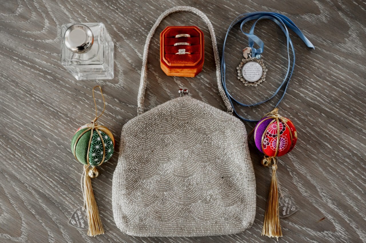 A pearl-beaded handbag with rings, perfume, two colorful ornaments, and a blue locket necklace lays on a gray surface as someone prepares for an Amaterra Winery wedding at The Heathman Hotel.