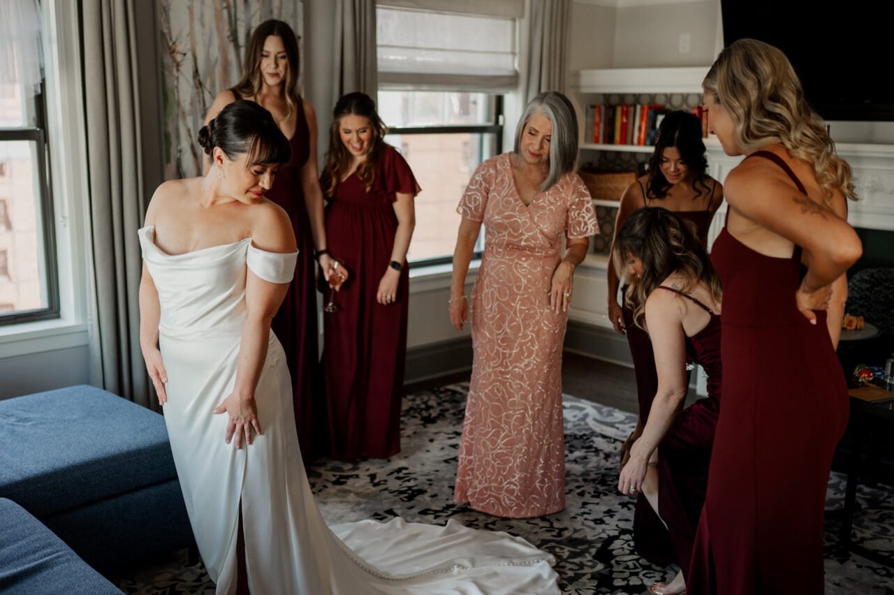 In a room bustling with excitement, the bride in her white gown stands gracefully as bridesmaids in red dresses surround her. A woman in a peach dress carefully adjusts the train, ensuring every detail is perfect while getting ready at The Heathman Hotel for the Amaterra Winery wedding.