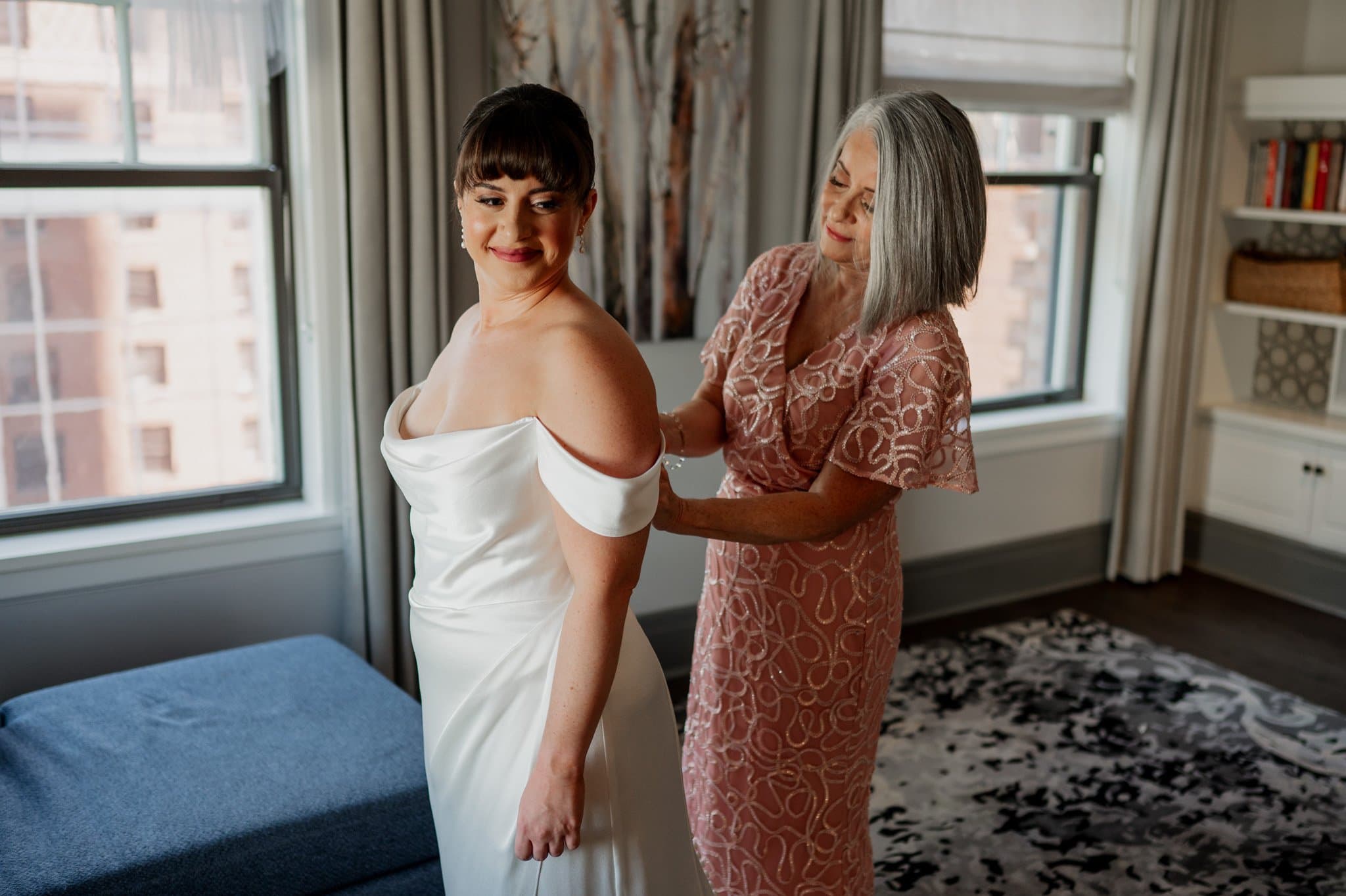 A woman in a white dress is getting ready at The Heathman Hotel, assisted by another woman in a pink dress, surrounded by the natural light streaming through large windows.