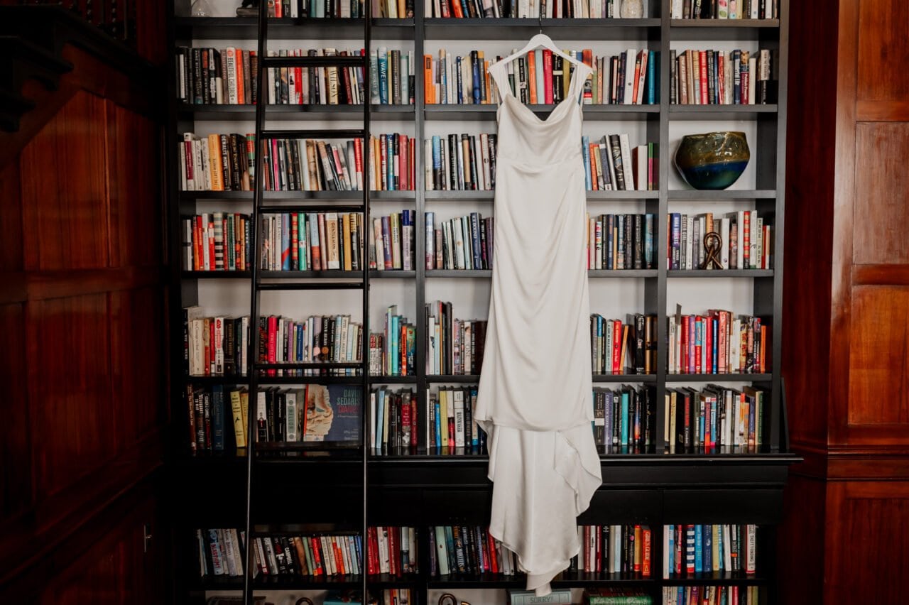 A white dress is elegantly draped on a bookshelf filled with books in a room featuring wooden walls and a ladder, creating an ambiance akin to getting ready at The Heathman Hotel for an Amaterra Winery wedding.
