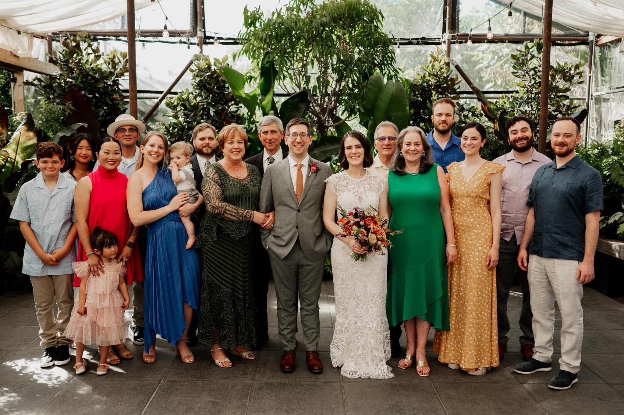 A diverse group of adults and children pose together in a lush greenhouse setting at Blockhouse, a Portland wedding venue, with a couple in elegant wedding attire.