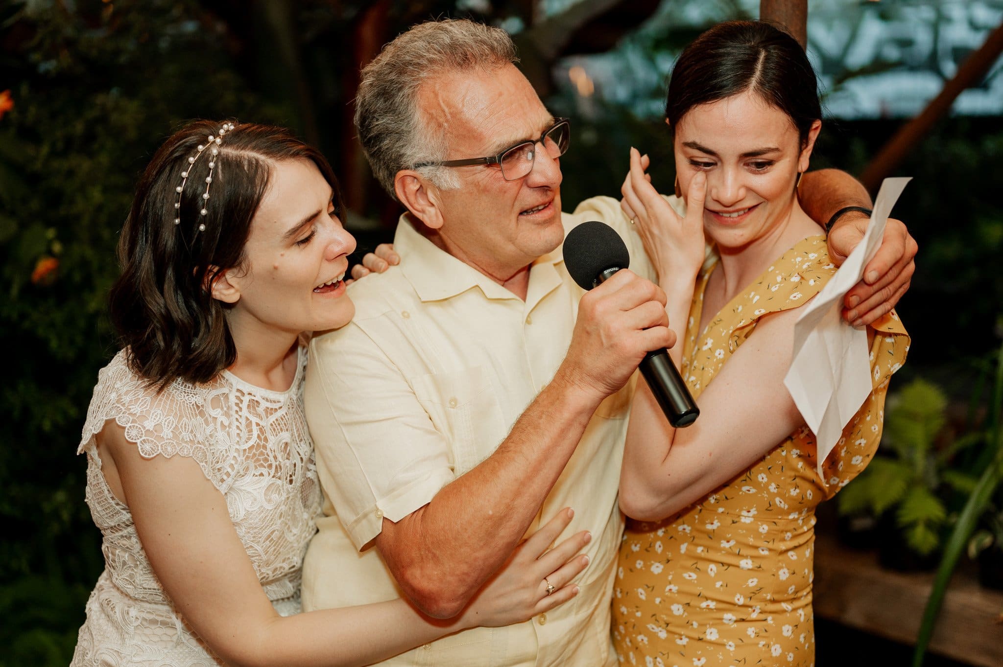 A man with a microphone hugs two smiling women at Blockhouse, a Portland wedding venue; one woman wears white lace while the other dazzles in a yellow floral dress, who wipes a tear from her eye.