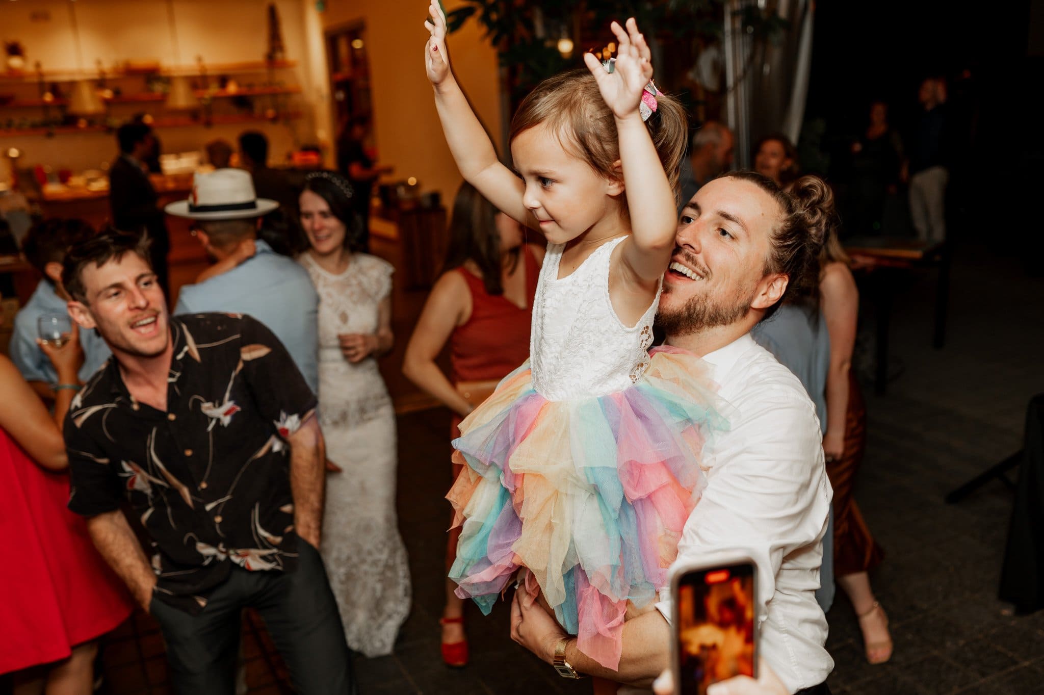 At a lively indoor party, a man joyfully lifts a smiling girl in a rainbow dress, capturing a moment fit for wedding photos at Blockhouse.