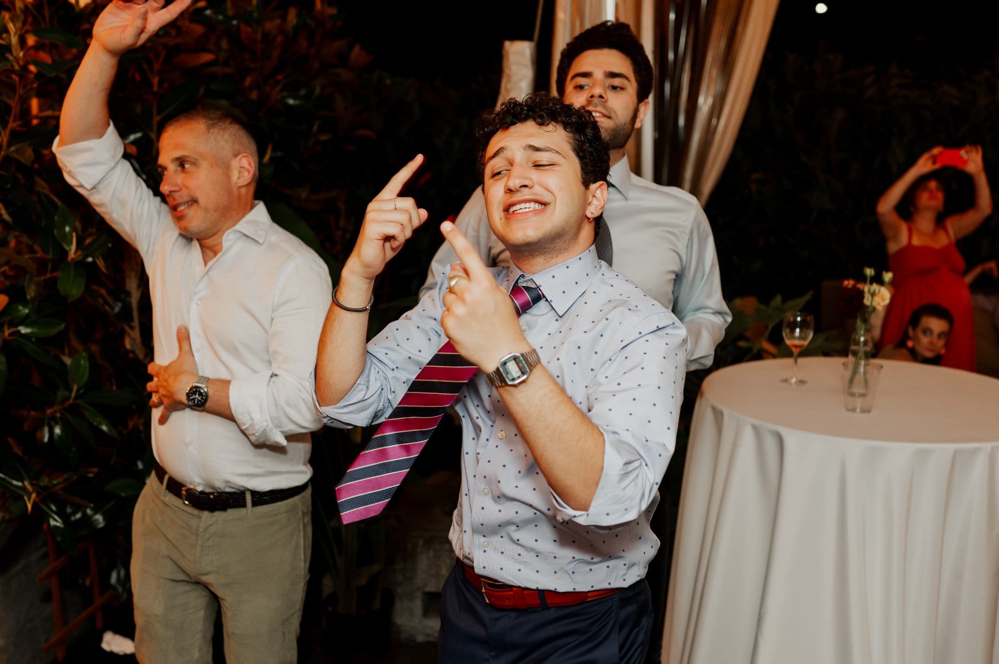 A person dancing joyfully at a party, wearing a striped tie and polka dot shirt, with others in the background. Its the perfect scene for wedding photos at Blockhouse, capturing pure happiness and celebration.