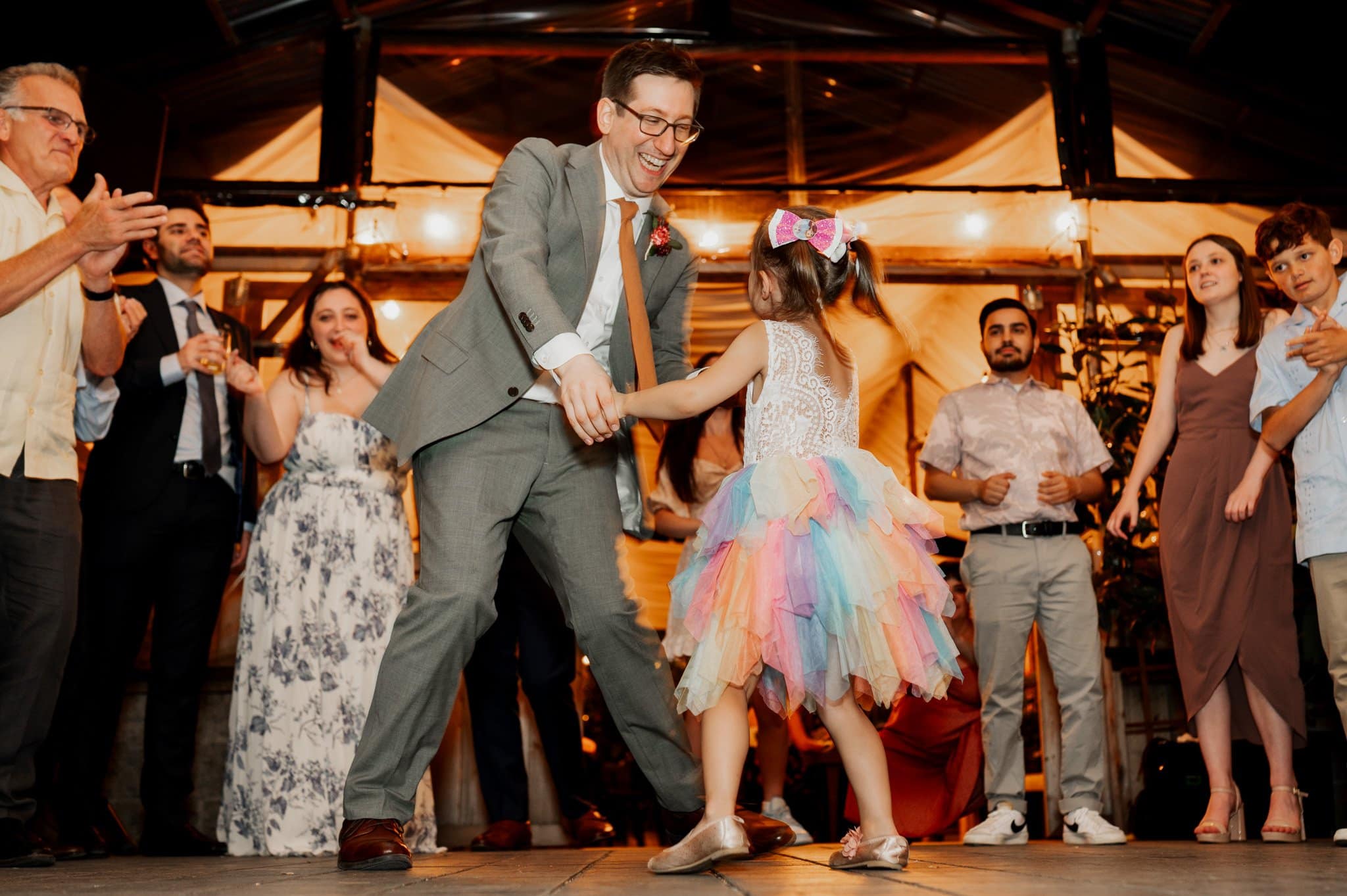 A man and girl dance joyfully on the dance floor, surrounded by smiling people in a festive celebration. The atmosphere is filled with laughter, capturing the essence of wedding photos at Blockhouse.