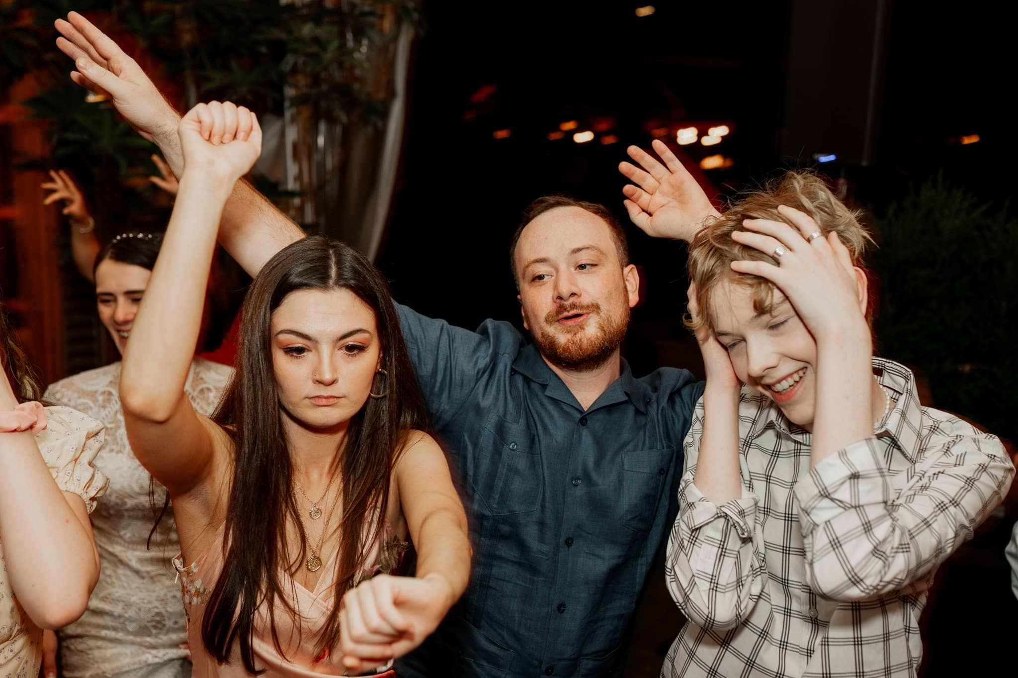 Three people are energetically dancing with arms raised, looking joyful and animated in the lively setting of a wedding at Blockhouse.