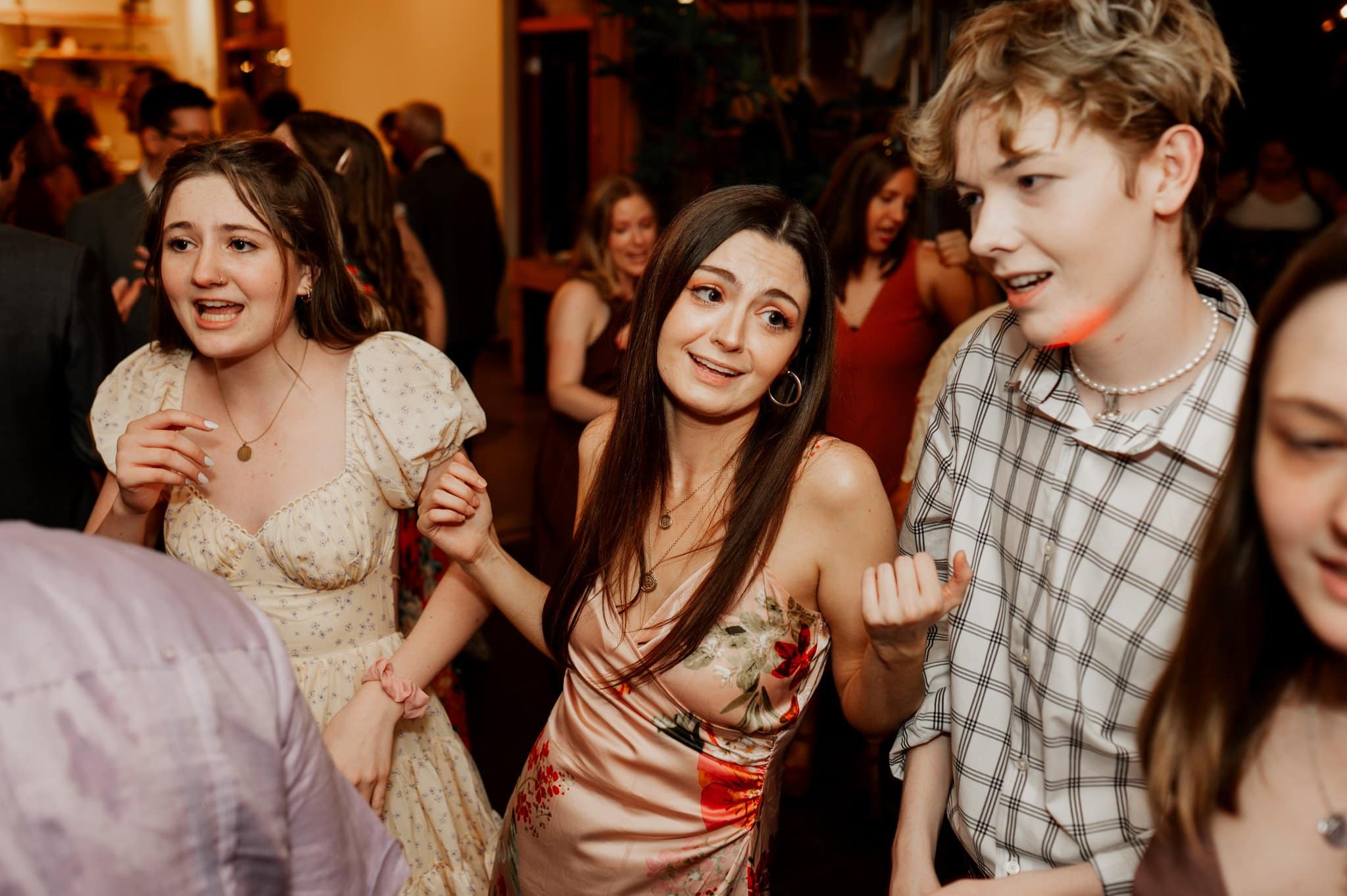 In a lively setting at the Blockhouse, three people are smiling and dancing in a group with others celebrating in the background, capturing the joy of wedding photos.
