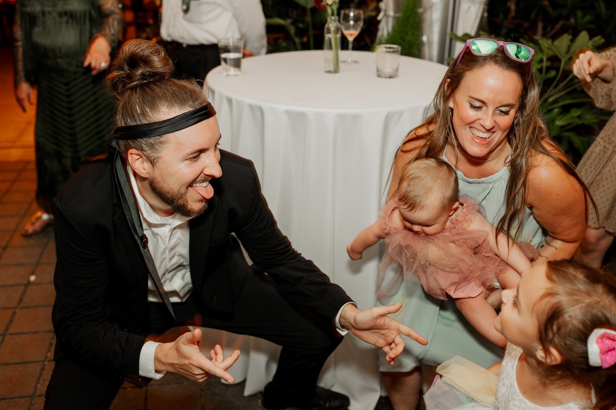 A man in a suit playfully interacts with a child while a woman holds a baby nearby at a lively wedding event. Captured as part of the wedding photos at Blockhouse, the scene buzzes with joy and memorable moments shared among family and friends.