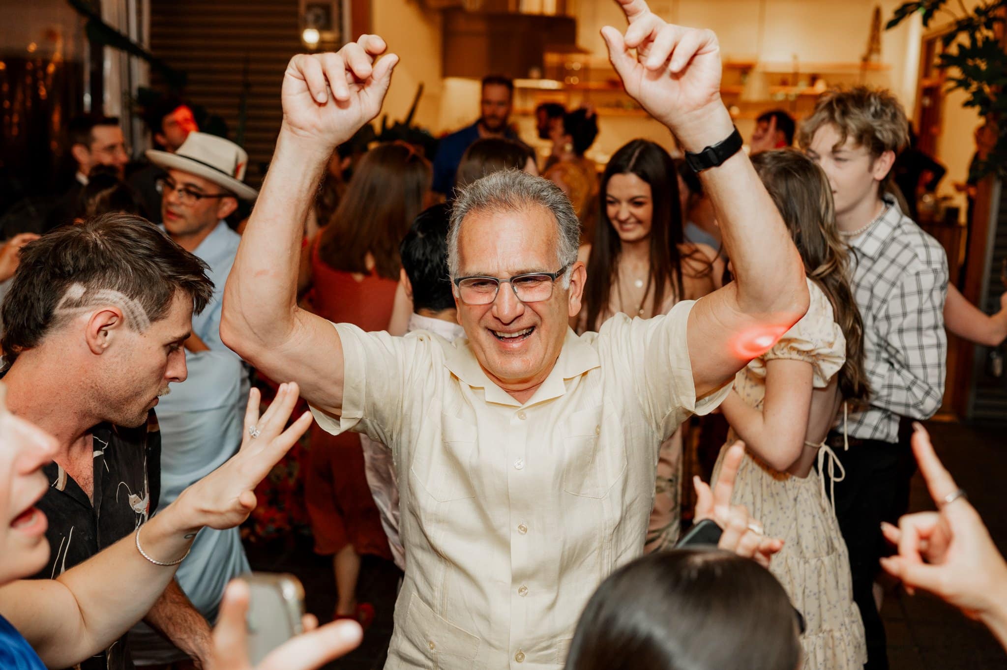 A person in glasses enjoys dancing at a lively party, surrounded by others with arms raised and smiling. Its a scene worthy of wedding photos captured at Blockhouse, where everyone revels in the cheerful atmosphere.
