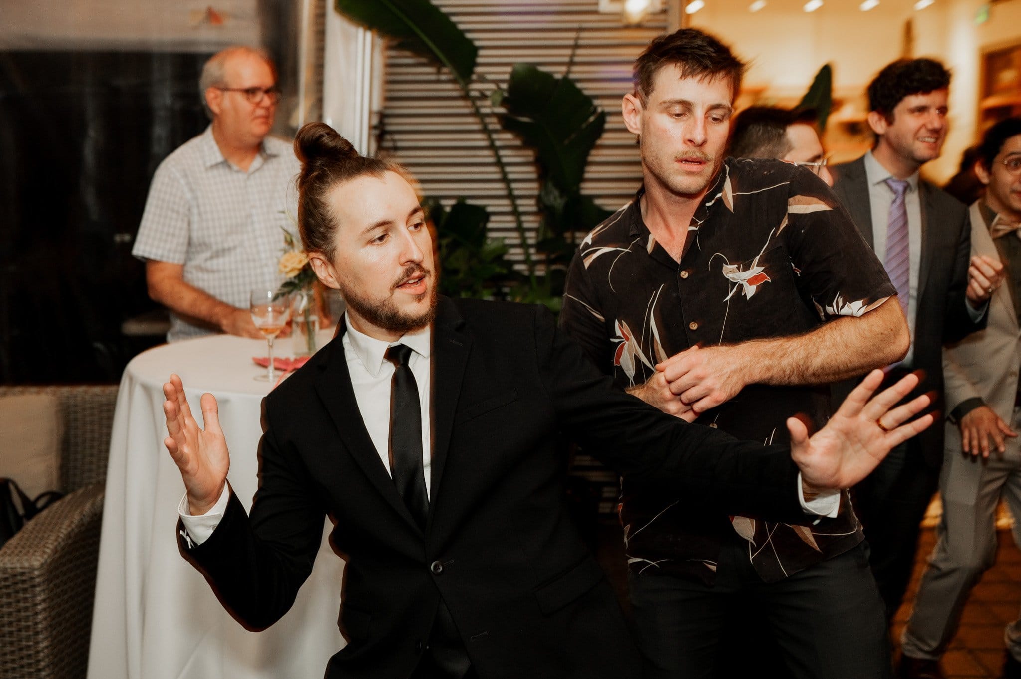 Two men are dancing energetically at an indoor event, surrounded by casually dressed guests and tables in the background. The lively atmosphere is captured perfectly in these wedding photos at Blockhouse.