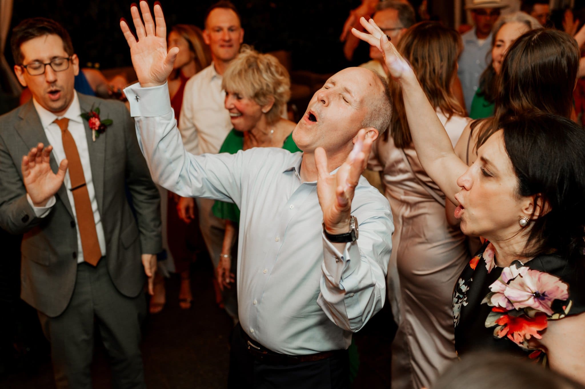 Group of people joyfully dancing and singing at a celebration, with some raising their hands in the air. The vibrant scene could easily be mistaken for wedding photos at Blockhouse, capturing the carefree spirit and excitement of the event.