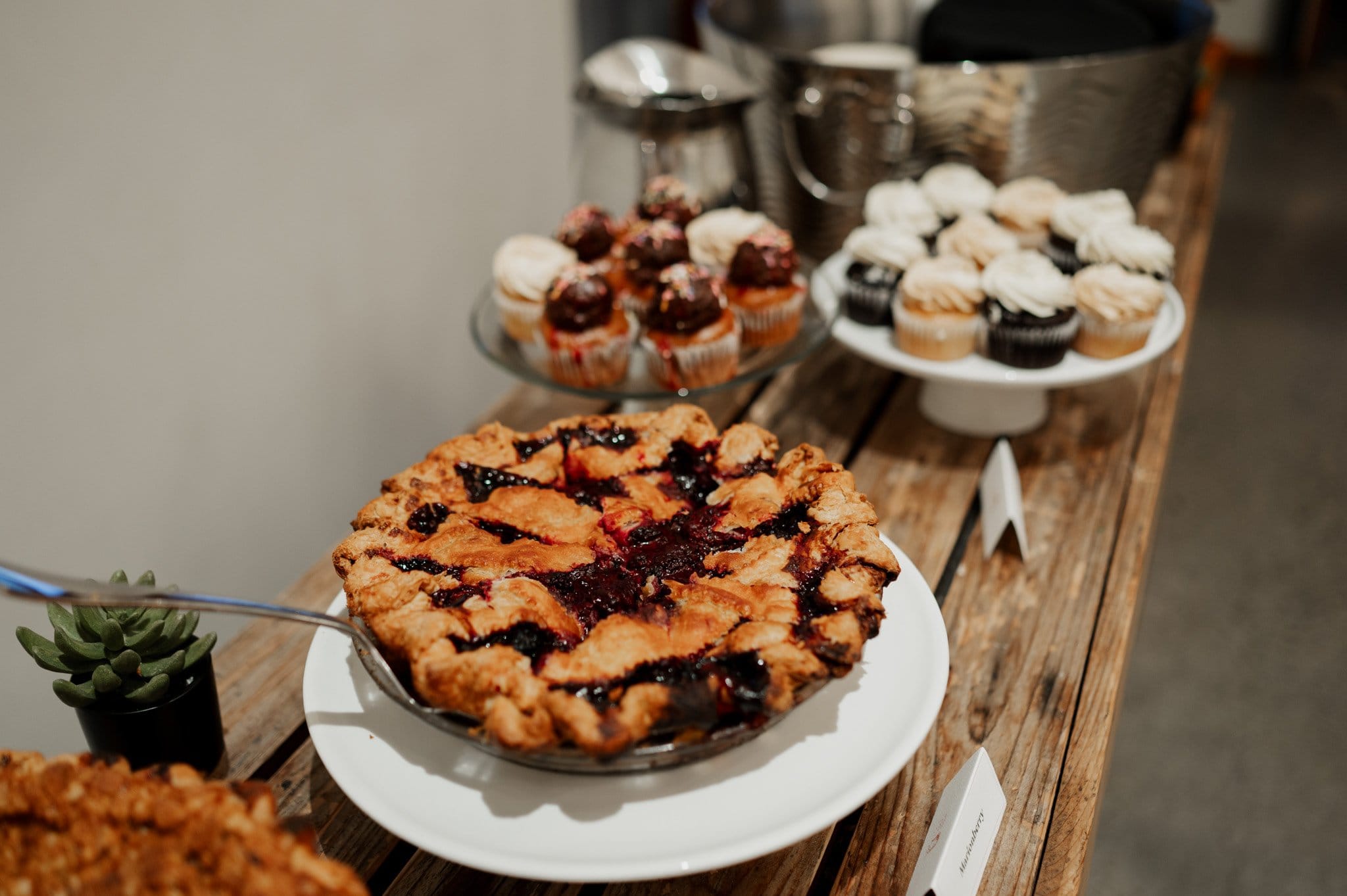 A berry pie sits elegantly on a wooden table with cupcakes and desserts in the background, adding a sweet charm reminiscent of wedding photos at Blockhouse.