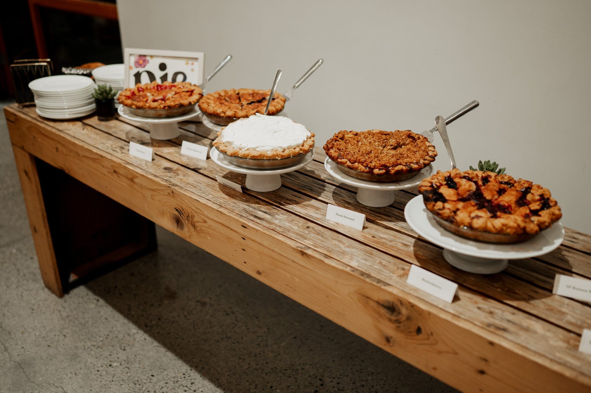 A wooden table displays five different pies on stands, each clearly labeled for guests. Plates are neatly stacked at one end, ready to be used. Ideal for adding a homely touch to your event, much like wedding photos at Blockhouse capture cherished memories in an intimate setting.