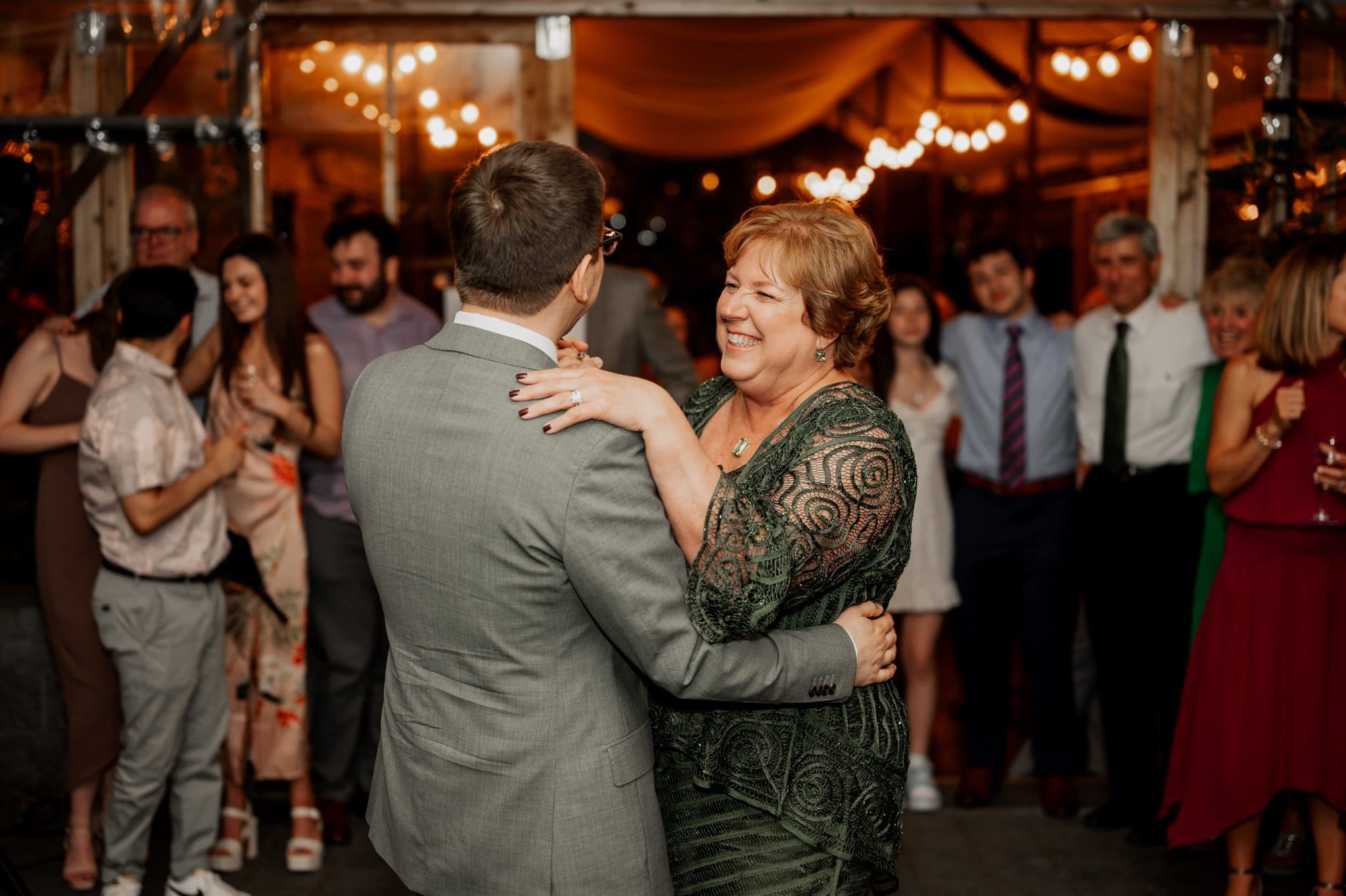 At a warmly lit party, a smiling woman in a green dress dances with a man in a suit, surrounded by people. The atmosphere is perfect for capturing wedding photos at Blockhouse.
