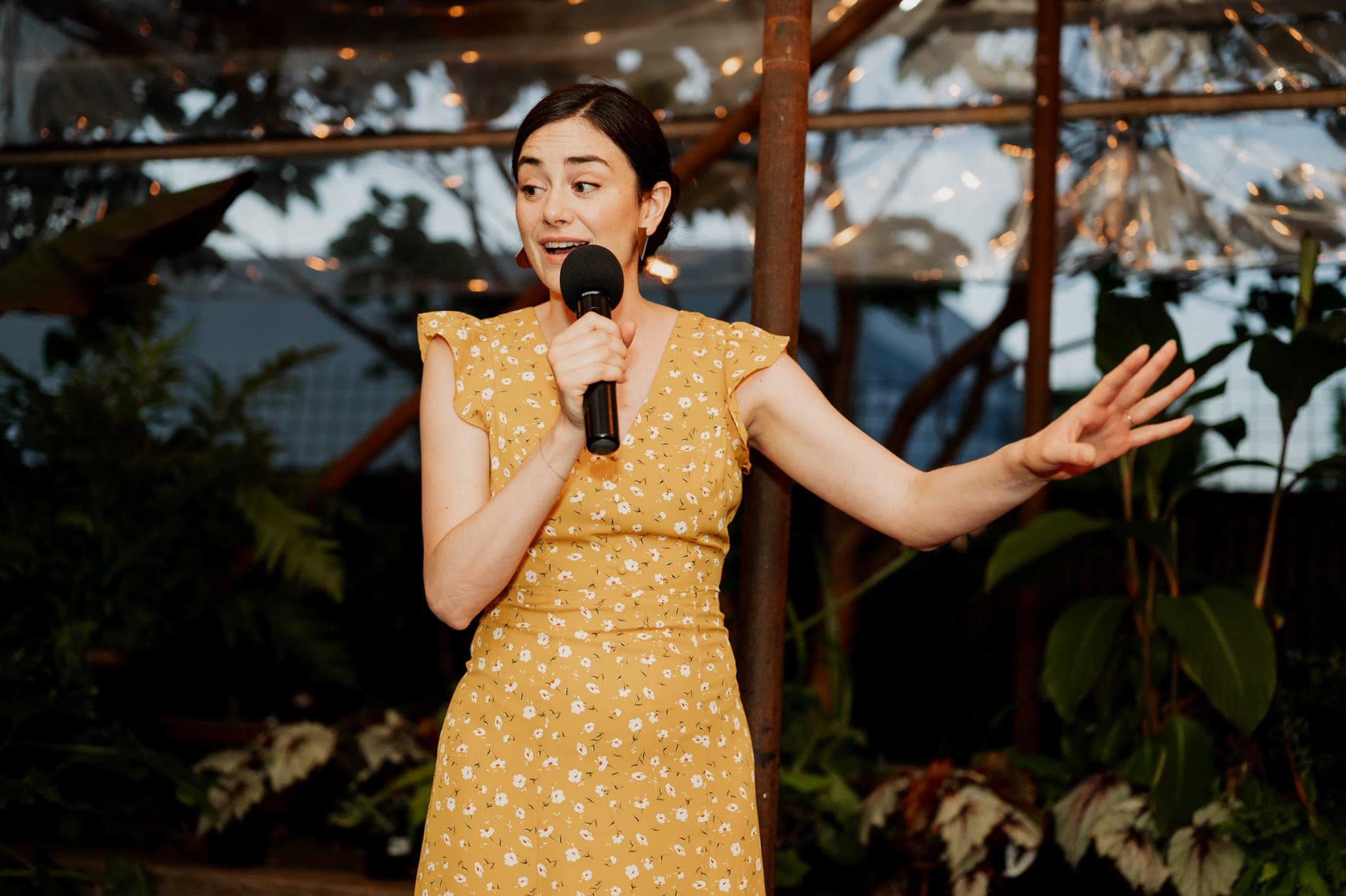 A person in a yellow dress speaks into a microphone, gesturing with one hand. The background features plants and lights, creating an atmosphere perfect for wedding photos at Blockhouse.
