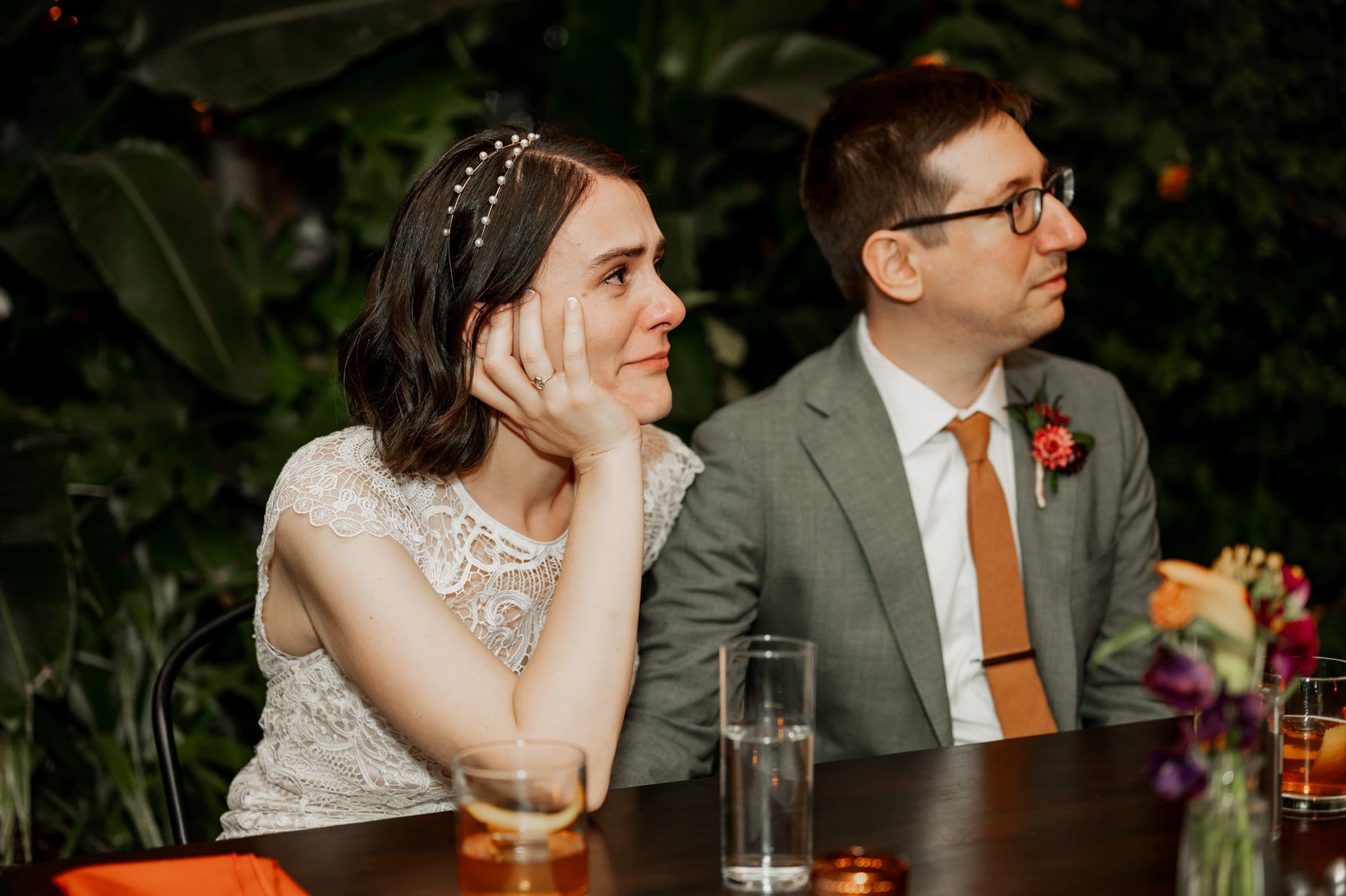 A couple sitting together at a table with drinks, looking thoughtful; the woman in a lace dress and the man in a gray suit. This intimate moment could easily be part of their wedding photos at Blockhouse, capturing a serene and meaningful connection.