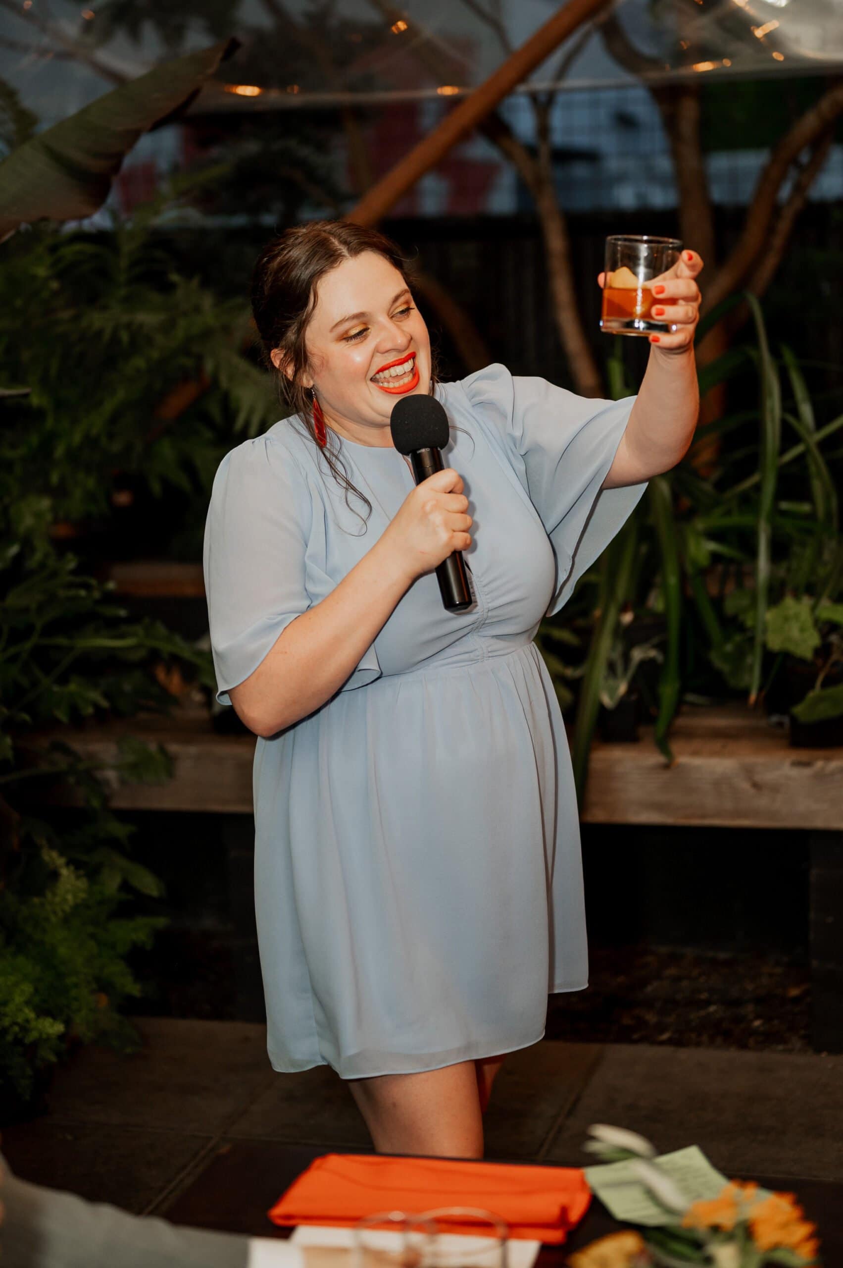 A person in a light blue dress holds a microphone and raises a drink, smiling amidst the lush greenery often seen in wedding photos at Blockhouse.