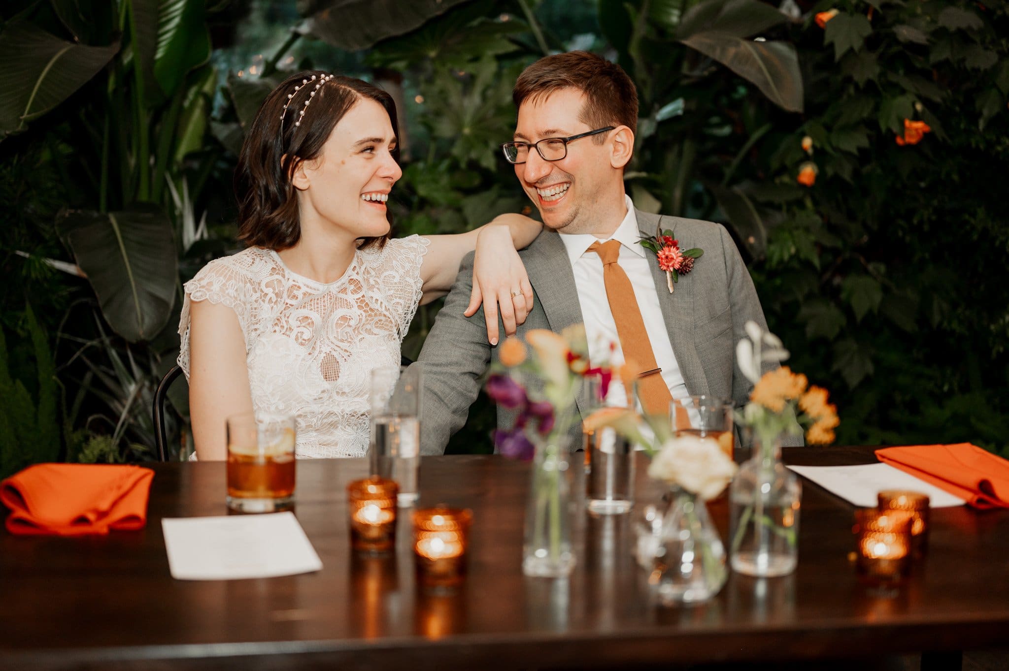 The smiling couple in wedding attire sits at a beautifully decorated table, surrounded by lush greenery, capturing their magical day with wedding photos at Blockhouse.
