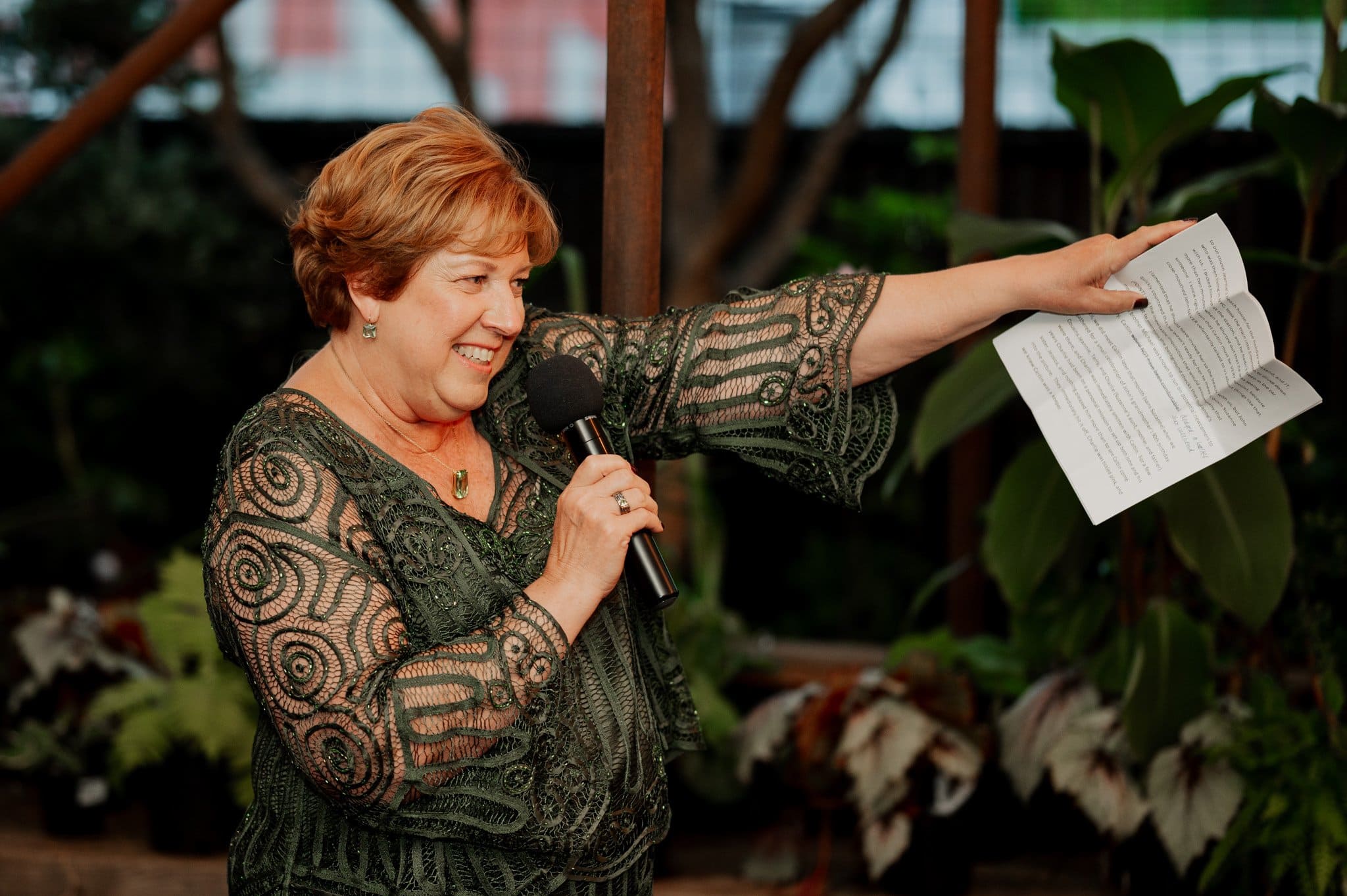 A woman in a green lace dress speaks into a microphone while holding papers, surrounded by lush plants. The setting, ideal for wedding photos at Blockhouse, adds a touch of natural elegance to the scene.