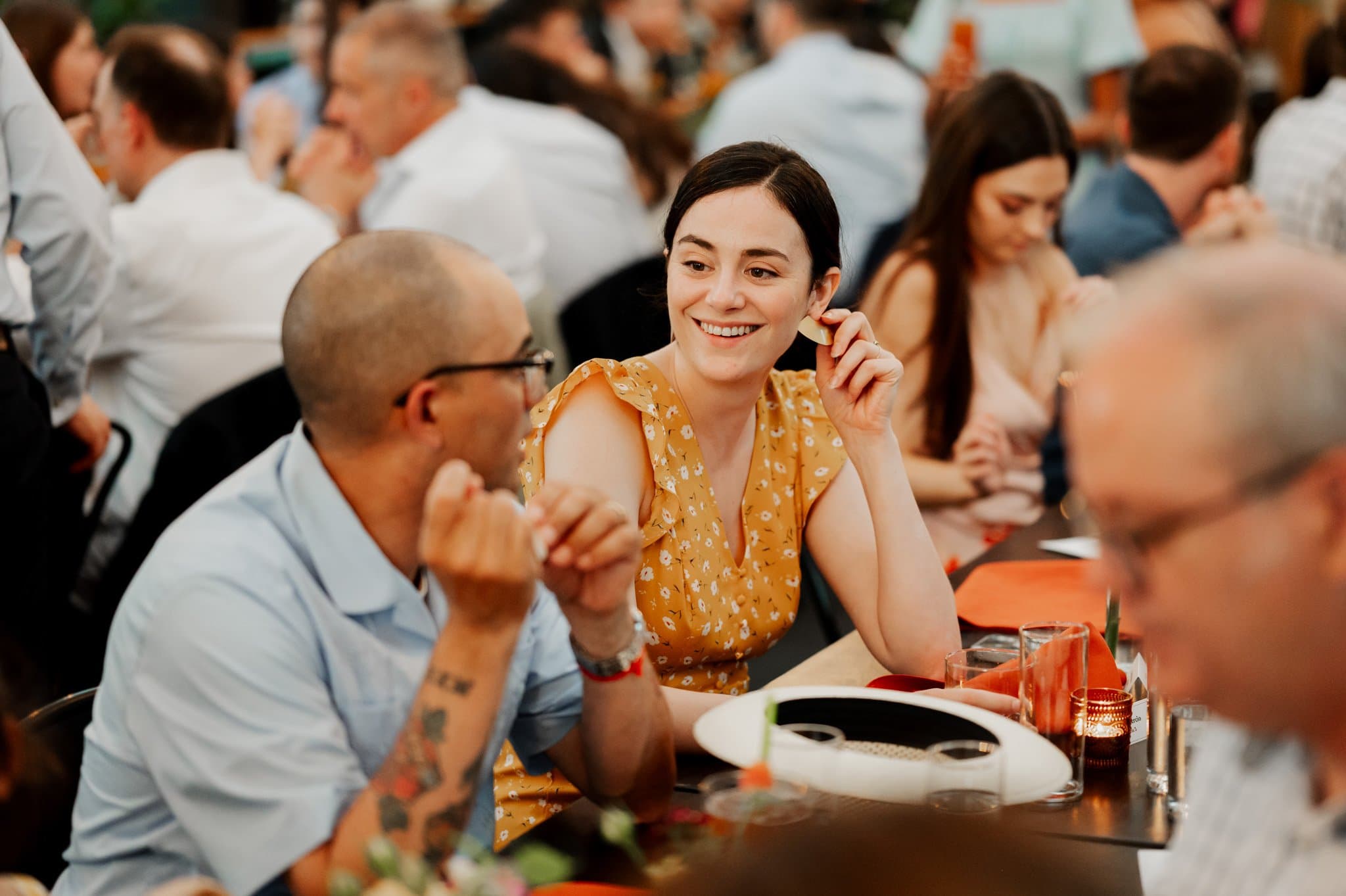 A smiling woman in a yellow dress sits at a crowded dining table at the Blockhouse, engaged in conversation with a man wearing glasses.