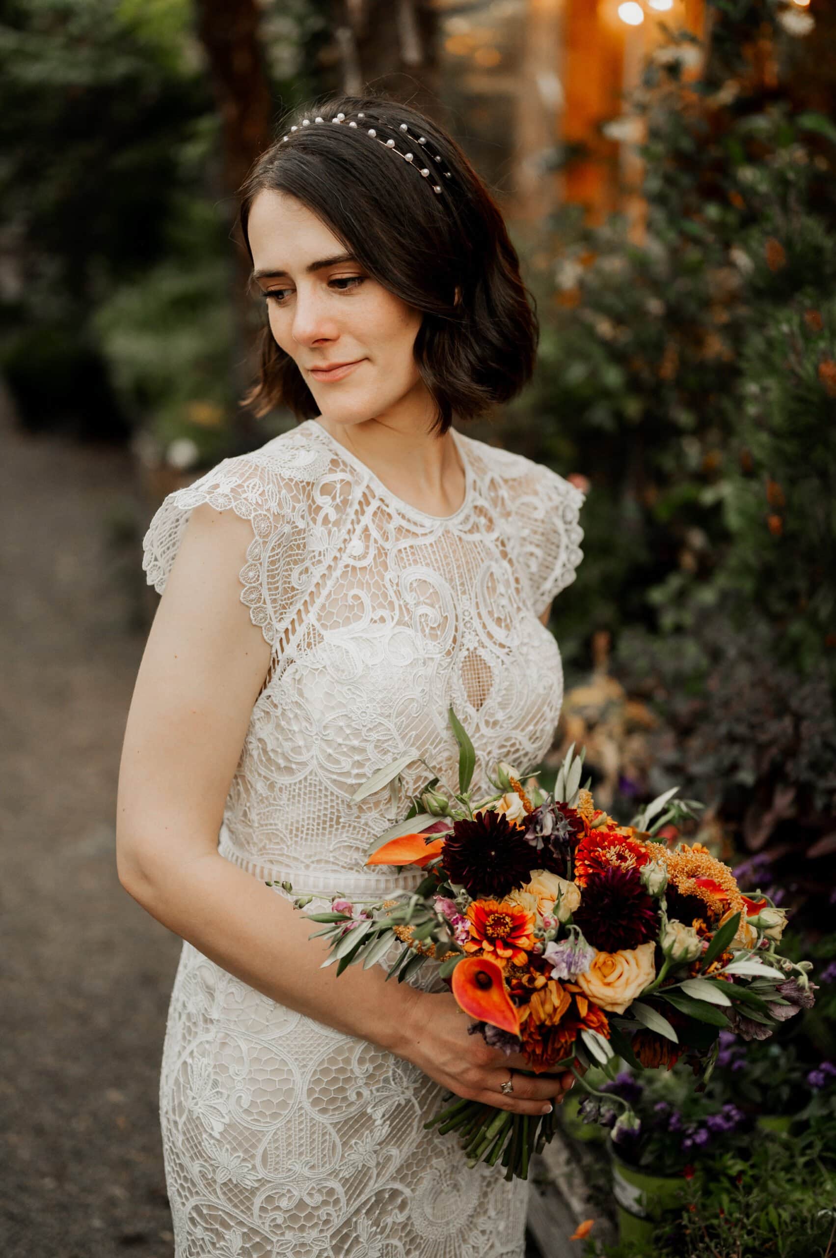 A bride in a lace dress stands elegantly outdoors, holding a vibrant bouquet of flowers. The picturesque setting is perfect for capturing wedding photos at Blockhouse, creating unforgettable memories of the special day.