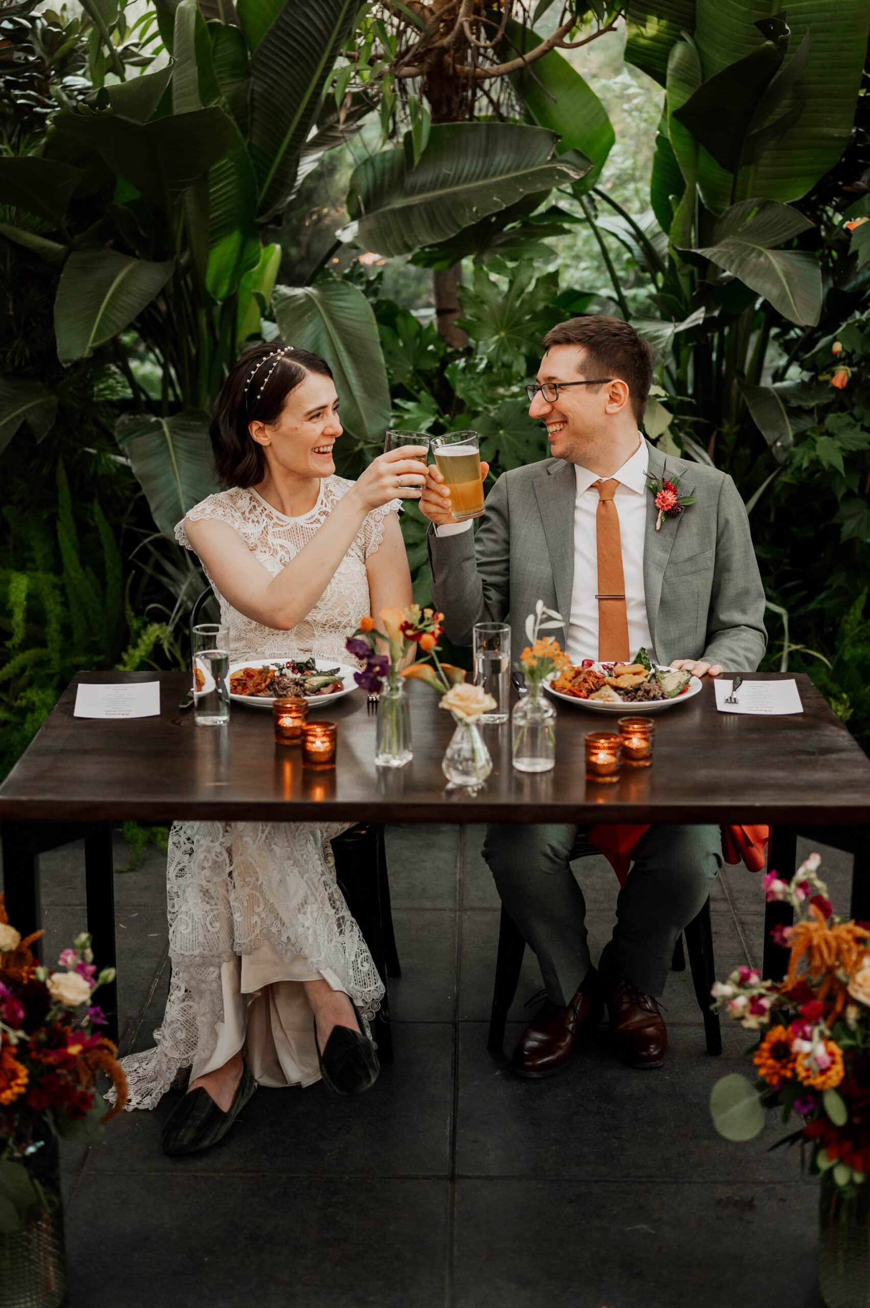 A couple in wedding attire toasts with drinks while sitting at a table surrounded by lush greenery and flowers, capturing a perfect moment for their wedding photos at Blockhouse.
