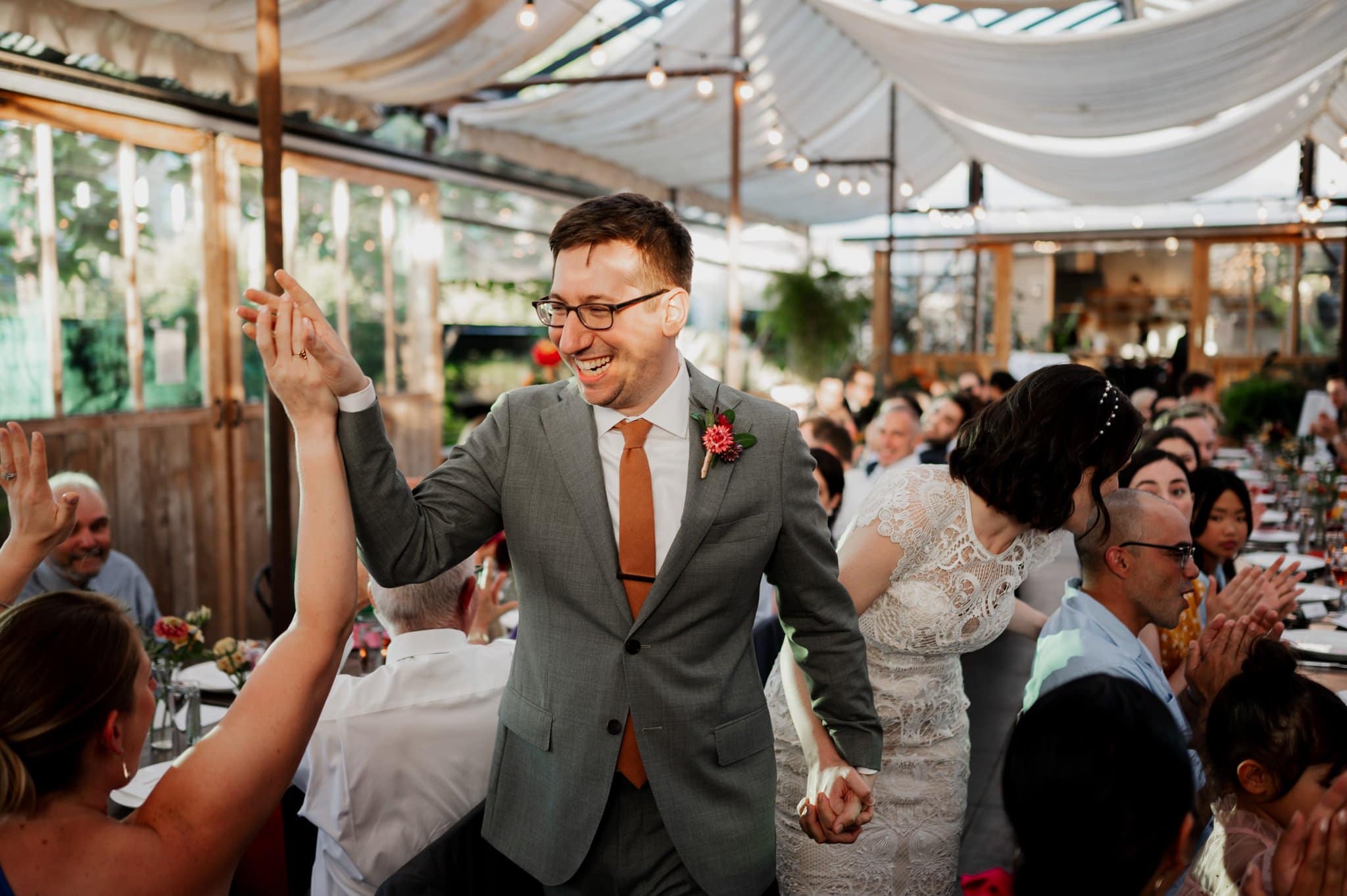 The groom in a suit high-fives a guest while walking hand-in-hand with the bride at a lively wedding reception, creating perfect wedding photos at Blockhouse.