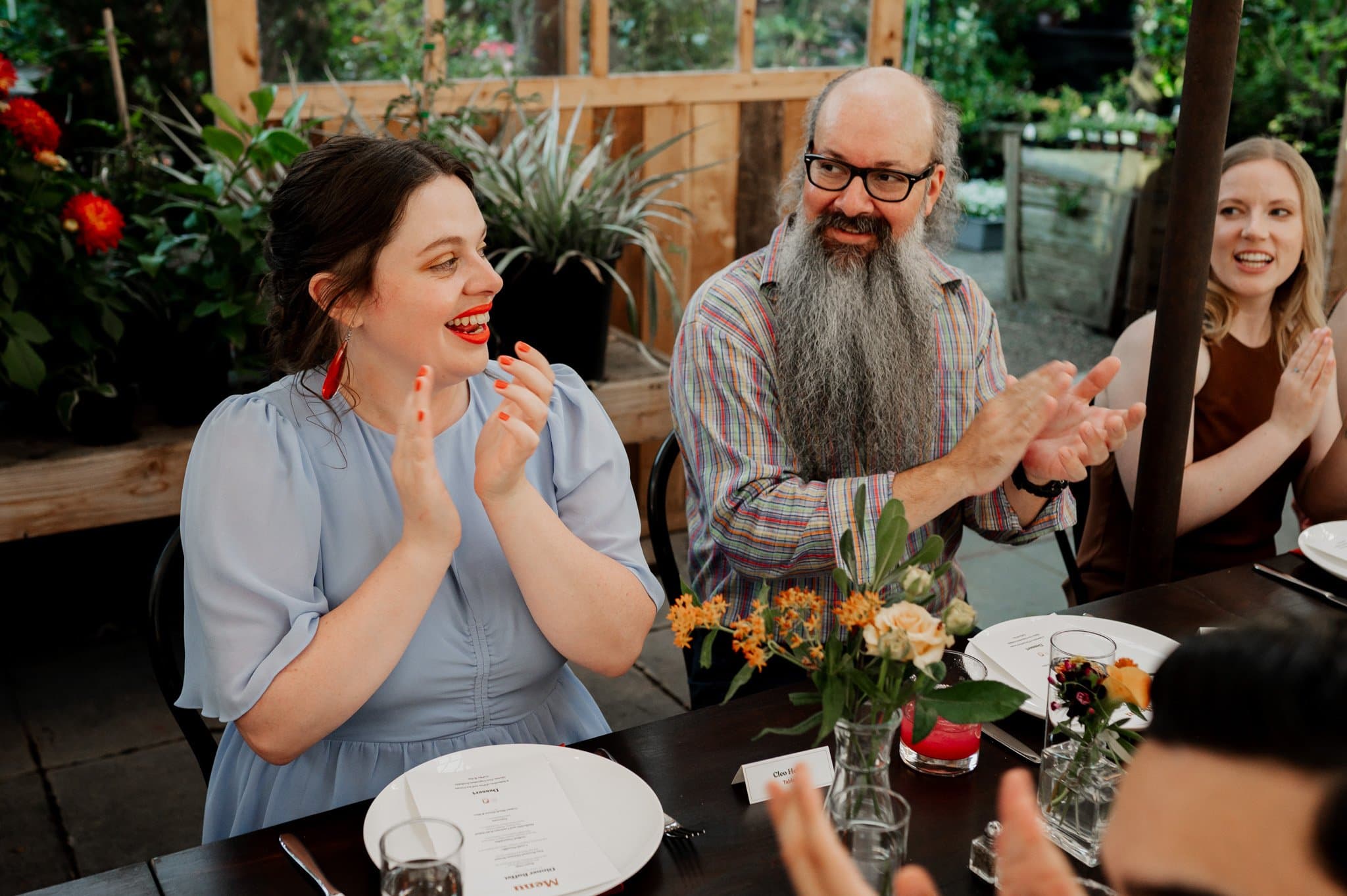 People clapping and smiling around a table adorned with flowers and menus in an outdoor setting, capturing the joyous atmosphere for wedding photos at Blockhouse.