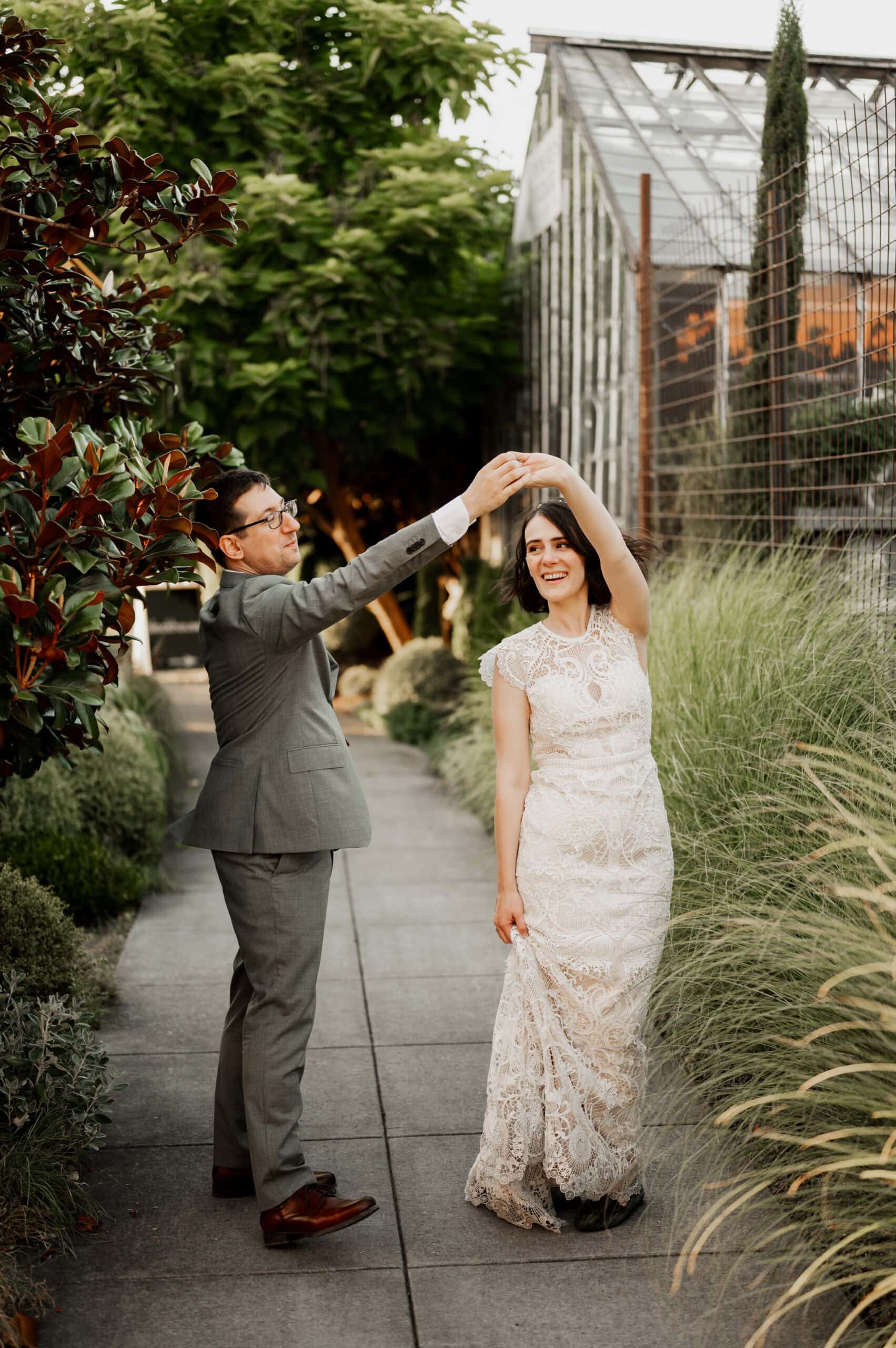 A couple dances along a garden pathway, with the groom in a gray suit and the bride in a lace gown, surrounded by vibrant greenery—a perfect setting for wedding photos at Blockhouse.