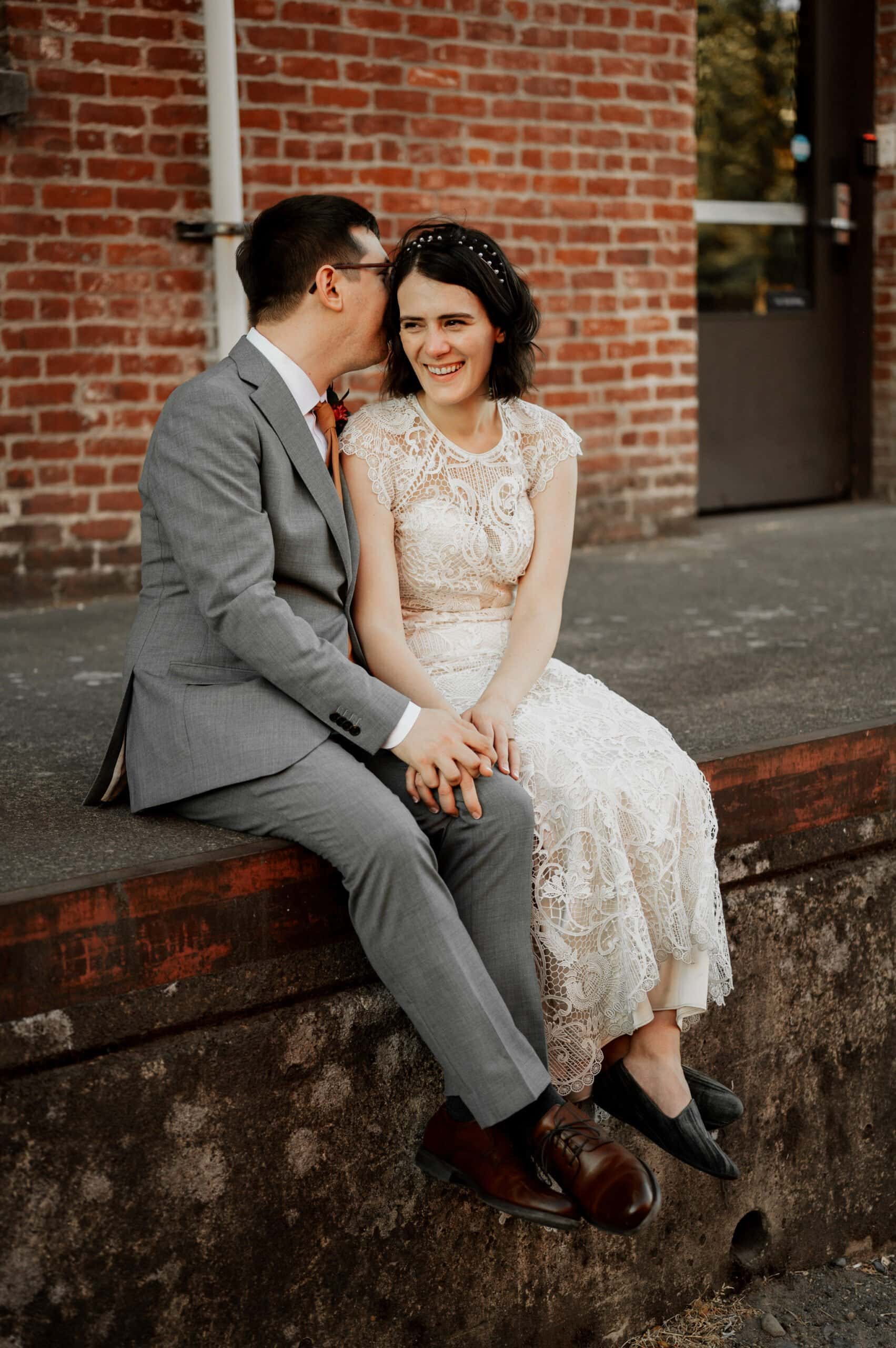 A couple in formal attire poses for wedding photos at Blockhouse, sitting on a ledge by a brick wall. The man leans in to whisper to the smiling woman as they hold hands.