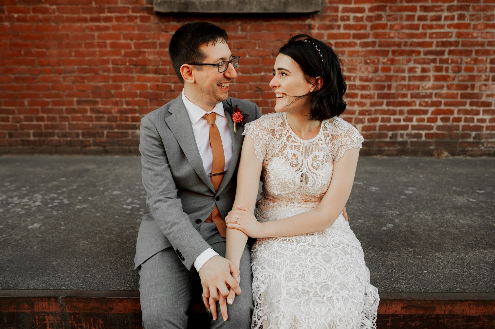 A couple in wedding attire sits on a ledge at Blockhouse, smiling at each other against a brick wall backdrop.