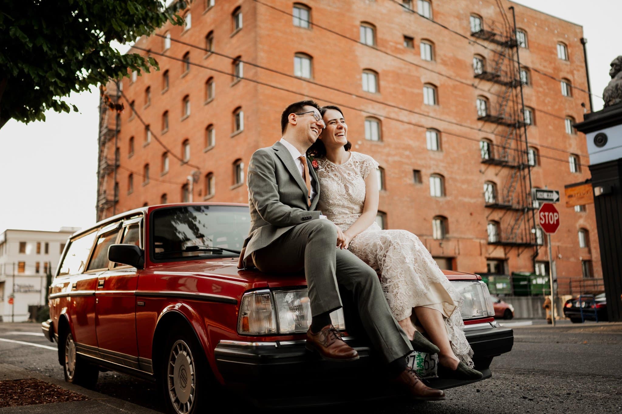 A couple in formal attire sits laughing on a red vintage car in front of a brick building, captured beautifully at Blockhouse during their wedding photos.