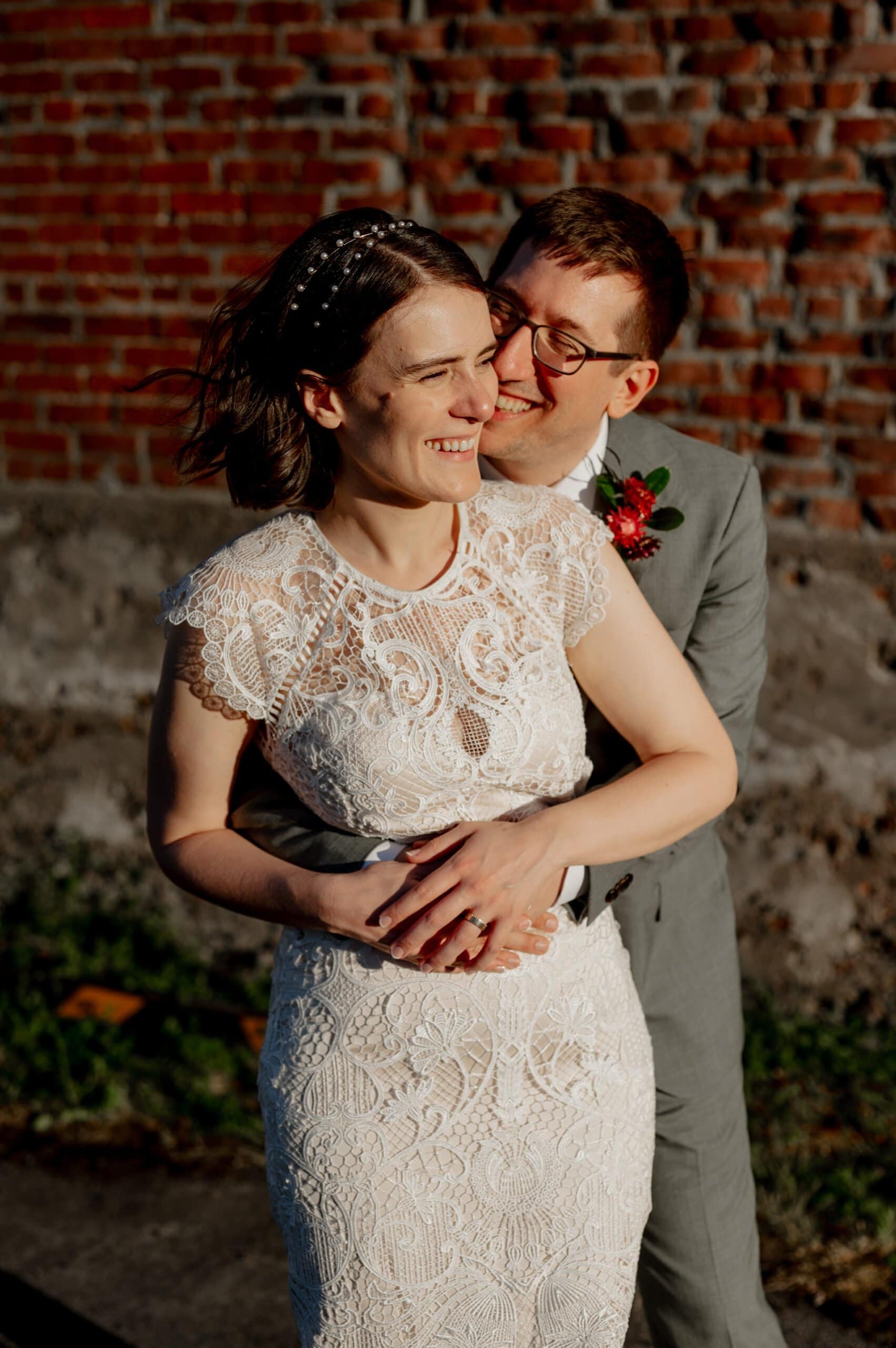 A couple in wedding attire lovingly embrace in warm sunlight, smiling against a brick wall as they capture their special day in wedding photos at Blockhouse.