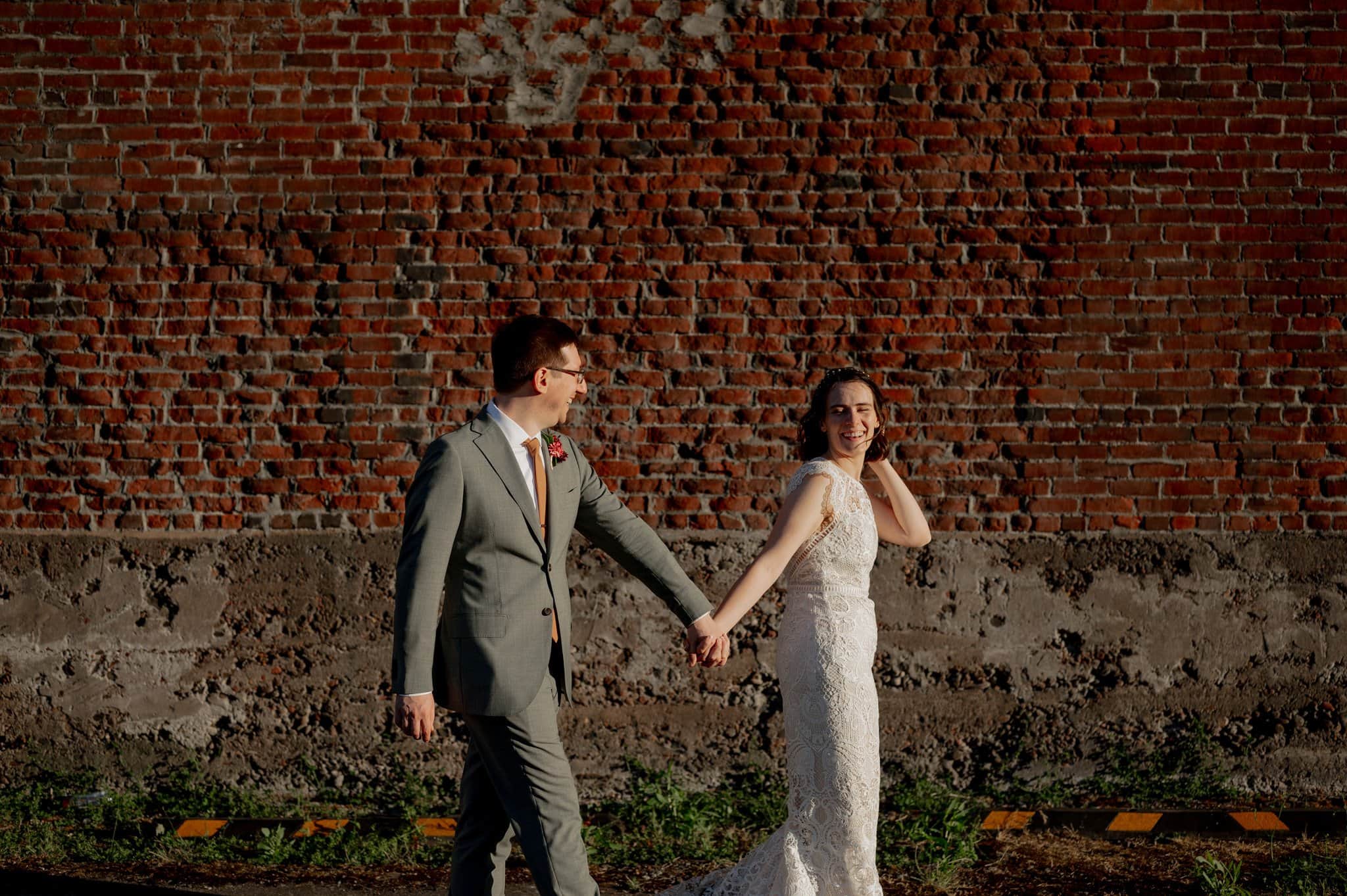 The couple in wedding attire joyfully hold hands while walking in front of a textured brick wall, capturing the perfect moment for their wedding photos at Blockhouse.