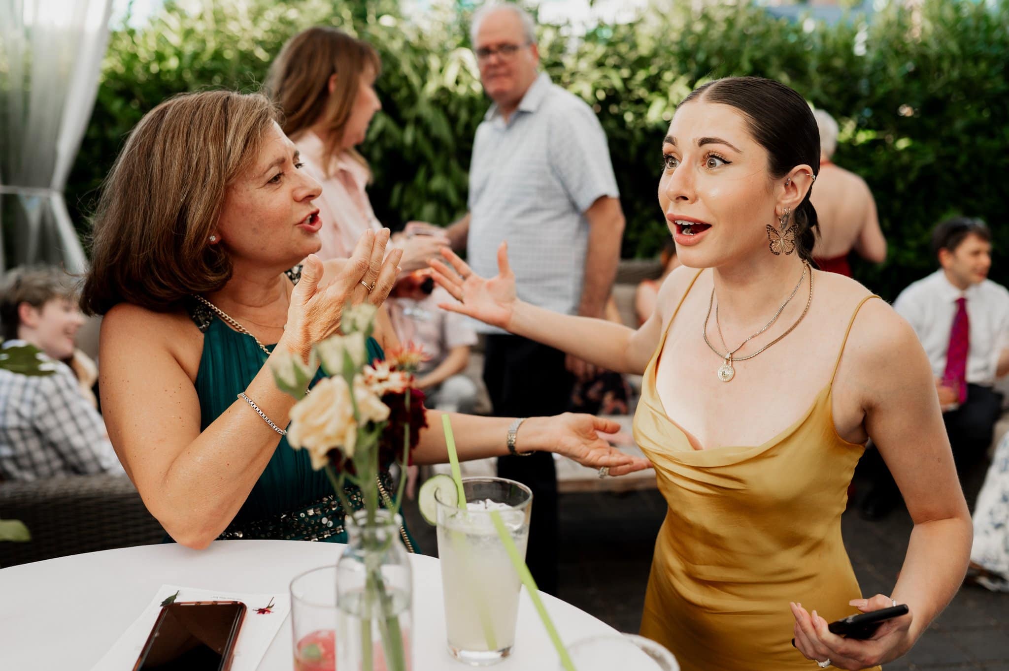 Two women animatedly conversing at an outdoor event, surrounded by others, with drinks on the table, capture a vibrant scene perfect for wedding photos at Blockhouse.