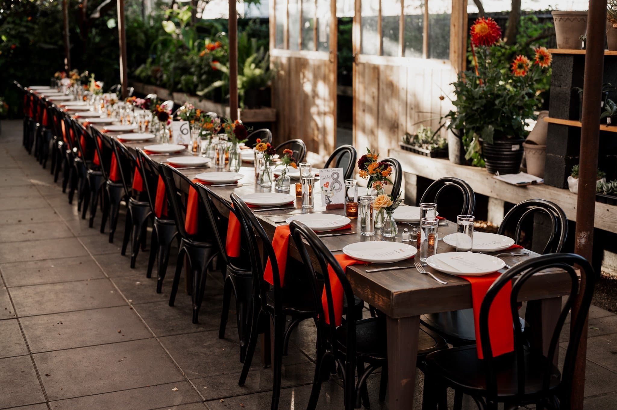 The long table is set for an outdoor event at Blockhouse, featuring black chairs, orange napkins, and stunning flower centerpieces. Perfect for capturing beautiful wedding photos against this elegant backdrop.