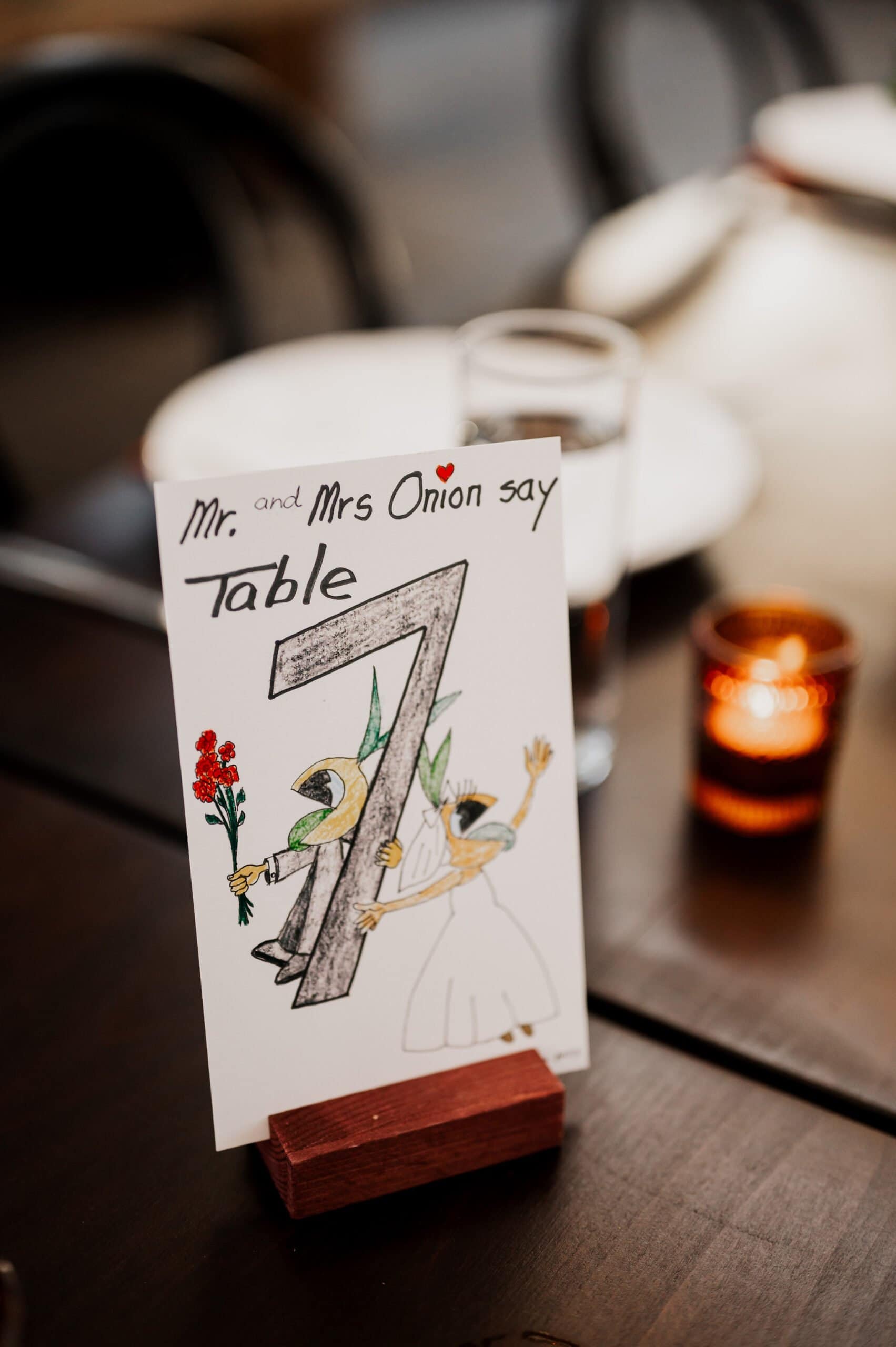 Card featuring cartoon onions dressed as bride and groom, holding a red flower, on a wooden stand. Perfect complement for wedding photos at Blockhouse.