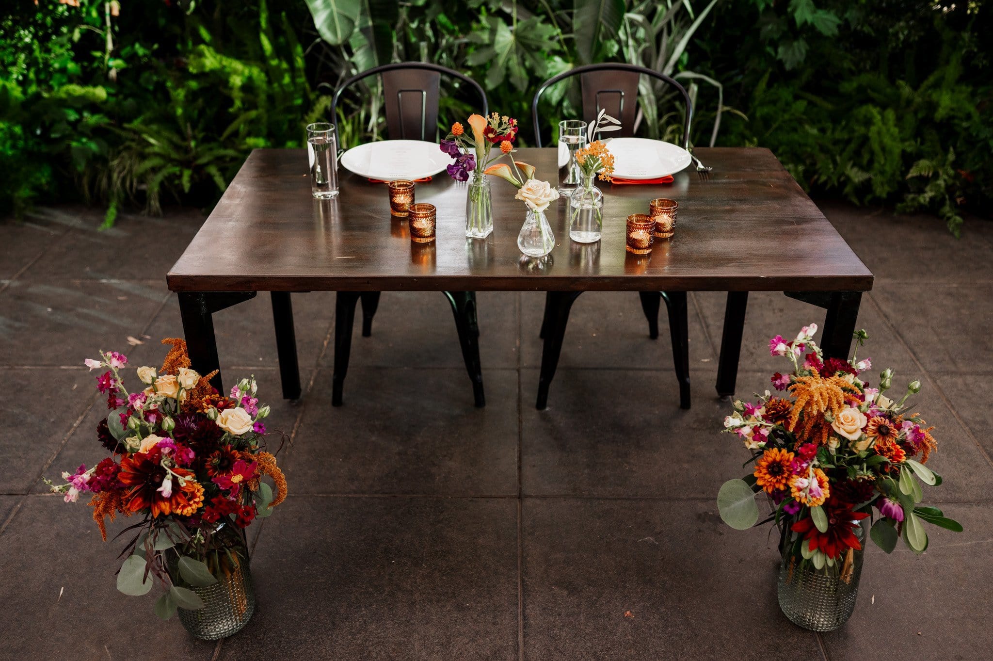 A wooden table set for two adorned with colorful floral arrangements and glassware, nestled amidst lush greenery, makes the perfect backdrop for wedding photos at Blockhouse.