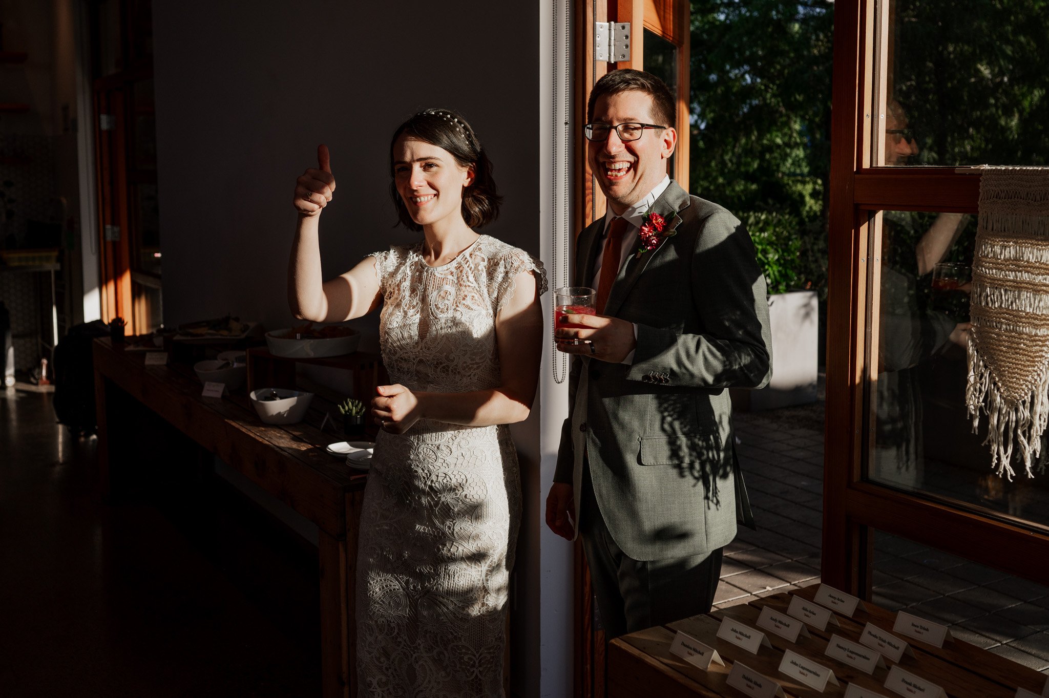A woman in a white dress gives a thumbs-up beside a man in a suit holding a drink, standing indoors near sunlight, capturing the joy of wedding photos at Blockhouse.