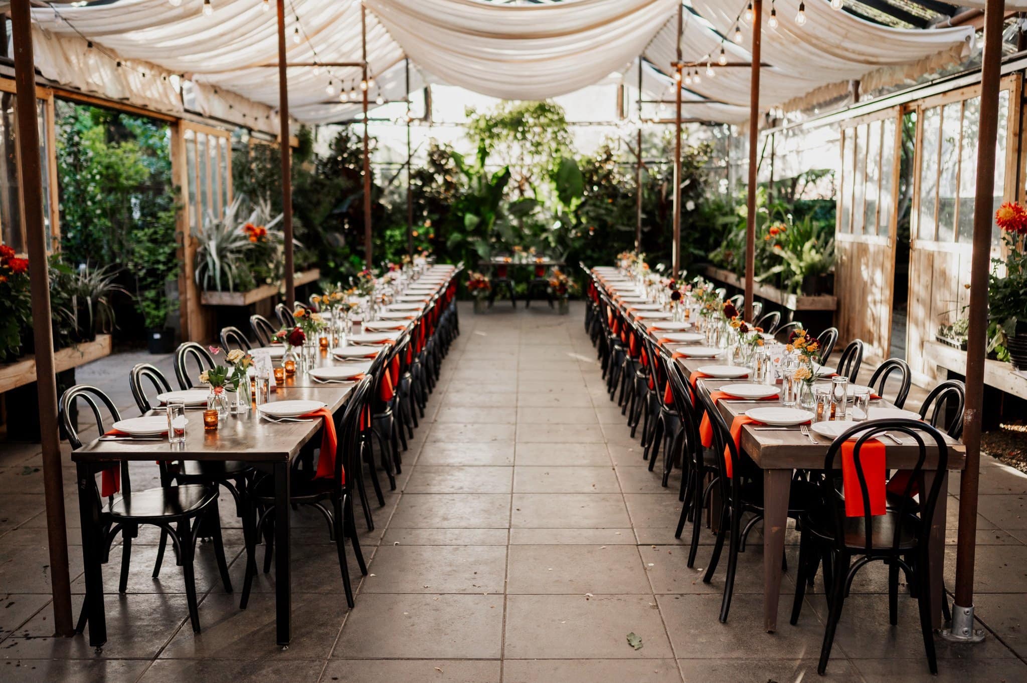 Experience an elegant outdoor dining setup perfect for wedding photos at Blockhouse. Featuring long tables, black chairs, red napkins, and floral centerpieces under gracefully draped fabric, this scenic arrangement offers a stunning backdrop for your special day.