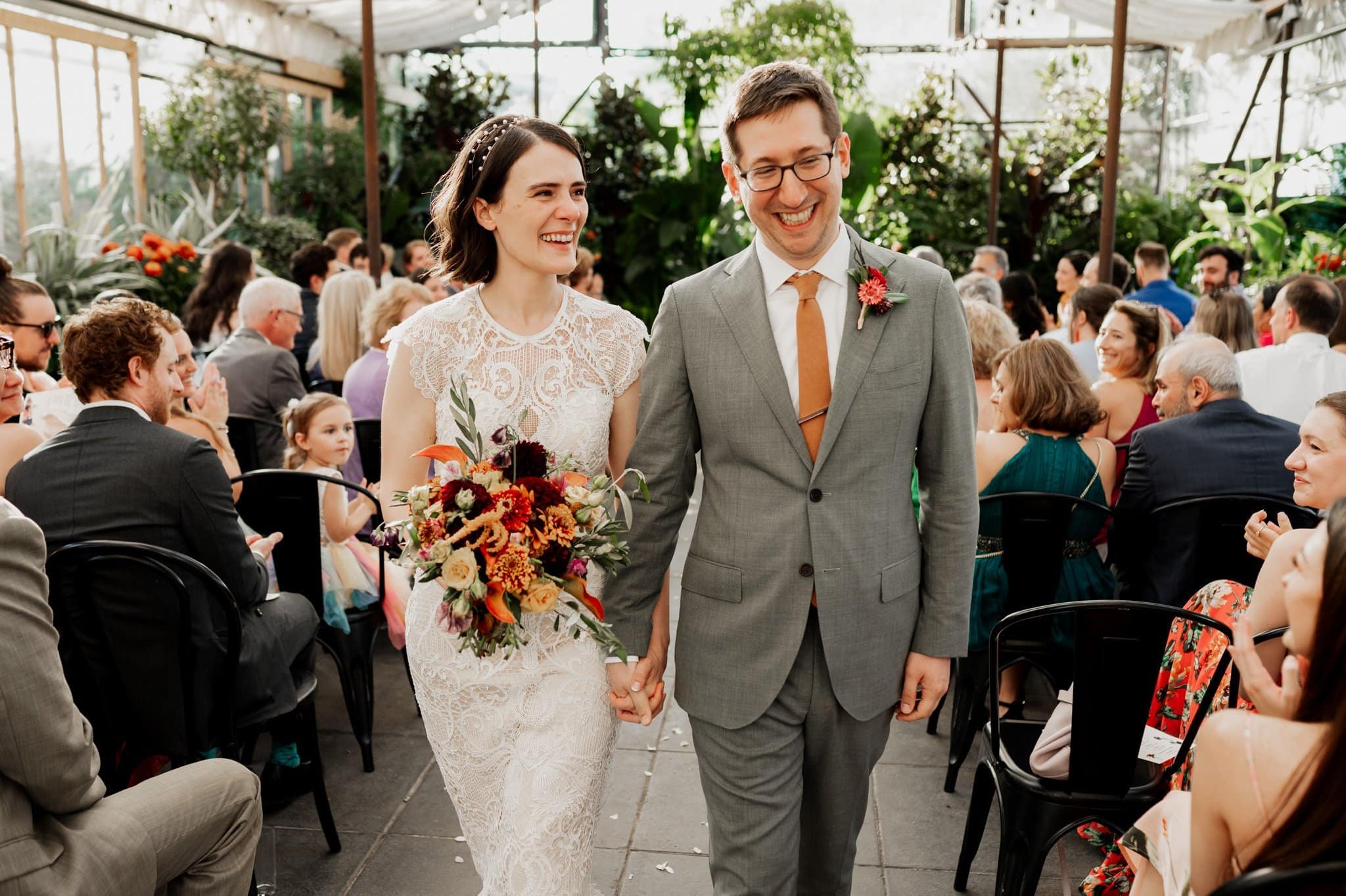 The bride and groom walk down the aisle, smiling in a greenhouse-style venue filled with guests. Their wedding photos at Blockhouse capture this joyful moment beautifully.