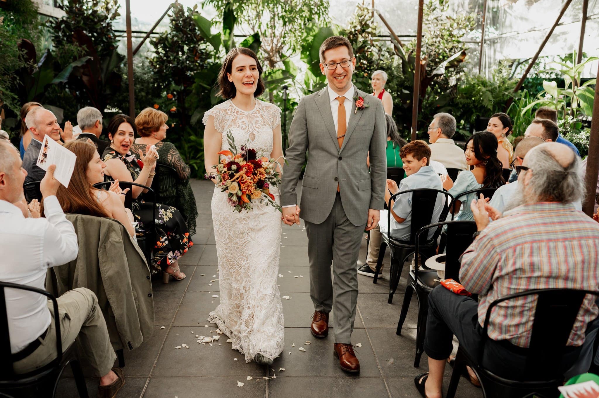 The bride and groom walk down the aisle, smiling as they are celebrated by applauding guests amid the lush greenery. Wedding photos at Blockhouse capture this beautiful moment in an enchanting setting.