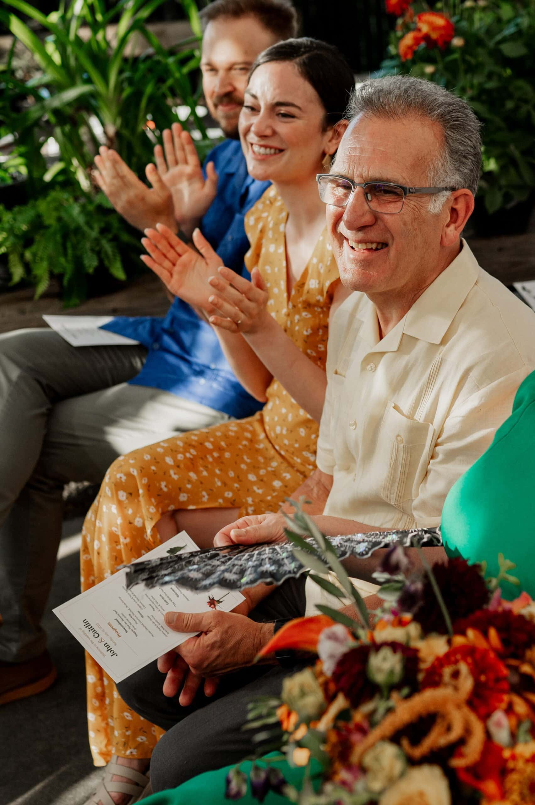 Attendees are sitting and clapping at the event, with flowers in the foreground and greenery in the background, capturing a moment akin to wedding photos at Blockhouse.