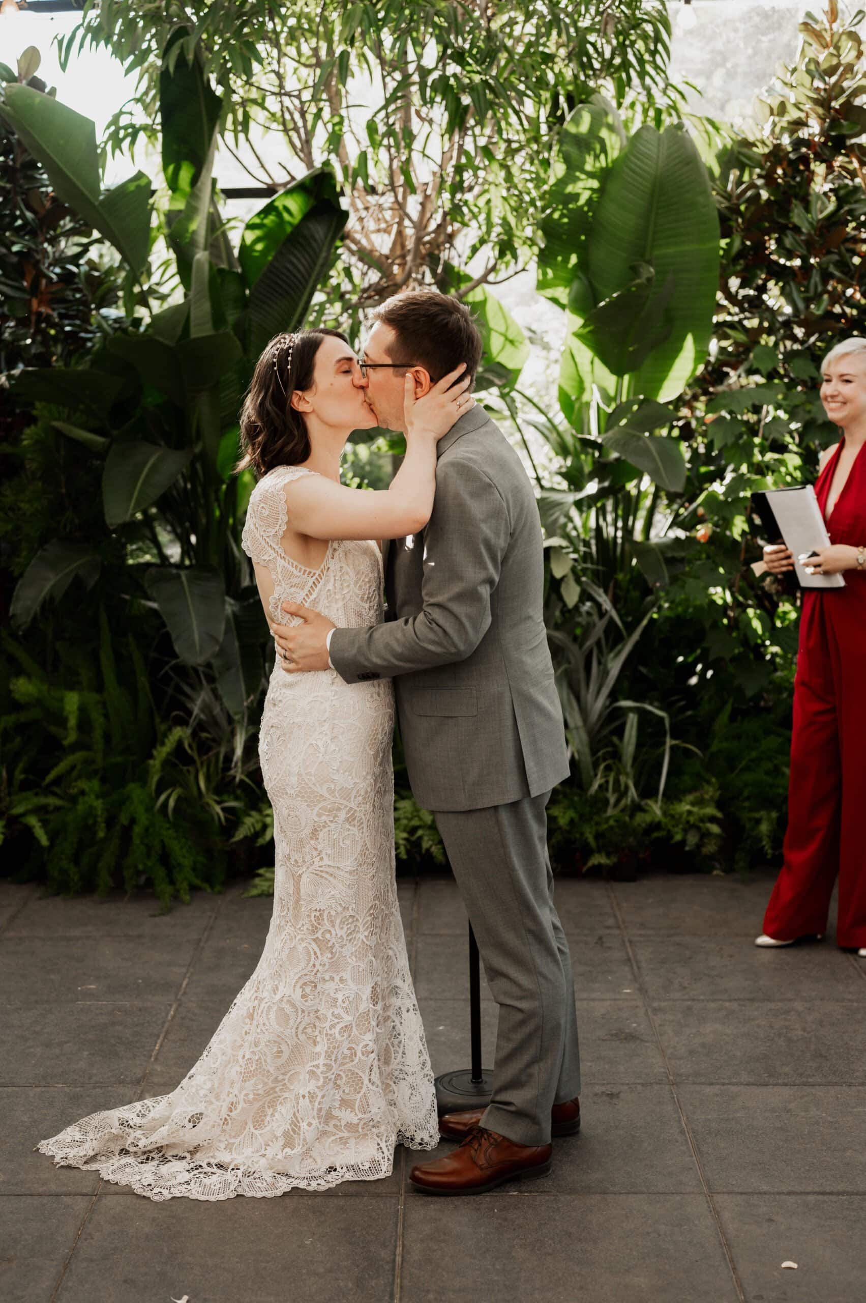 A couple shares a kiss during their wedding ceremony in a lush, leafy garden setting, perfect for wedding photos at Blockhouse. Nearby, a person in red stands by as the scene unfolds.