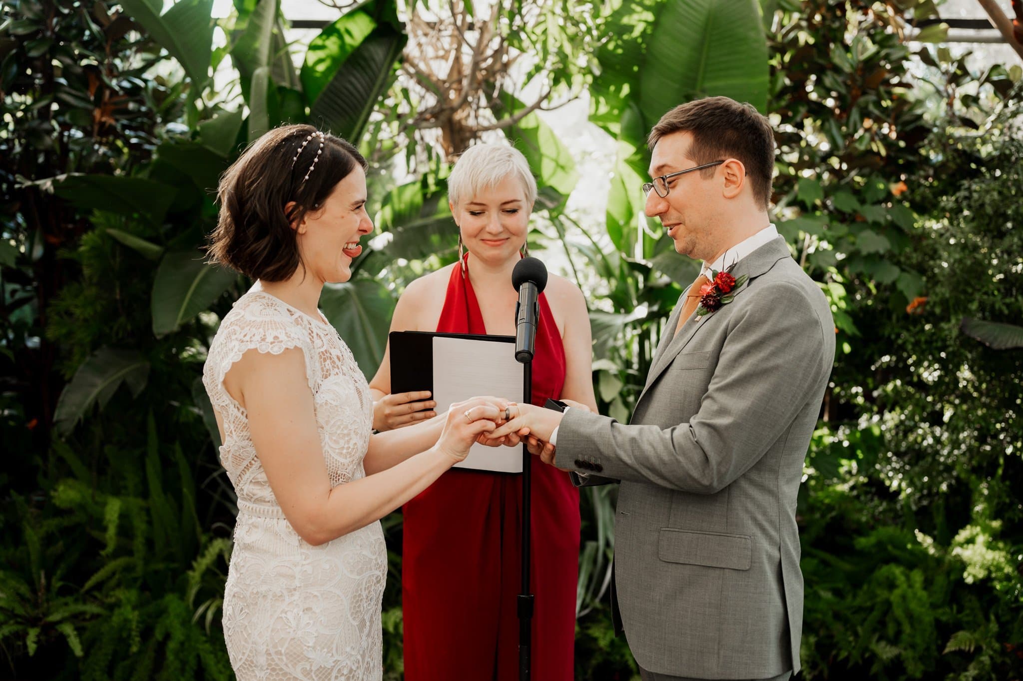 A bride and groom exchange rings during a wedding ceremony at Blockhouse, with an officiant in a red dress, surrounded by lush greenery.