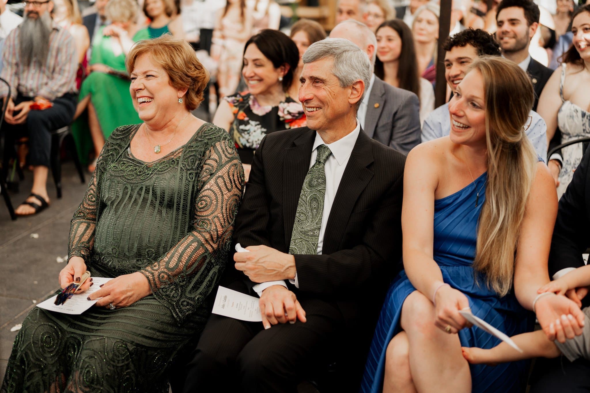 A group of people seated at a wedding at Blockhouse, smiling and laughing together, with a woman in a blue dress on the right.