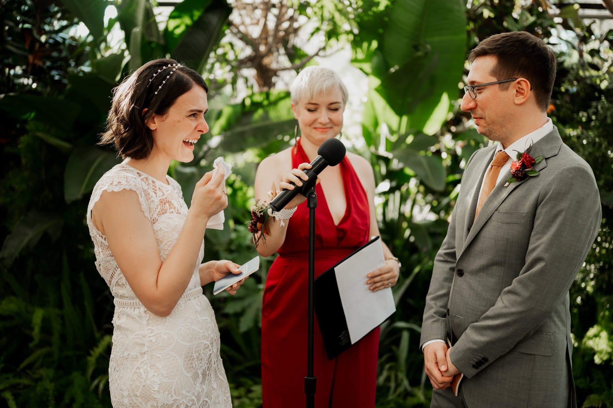 Bride laughing during vows, groom smiling warmly as the officiant holds the microphone. Lush green foliage provides a stunning backdrop, capturing the joy of their wedding photos at Blockhouse.