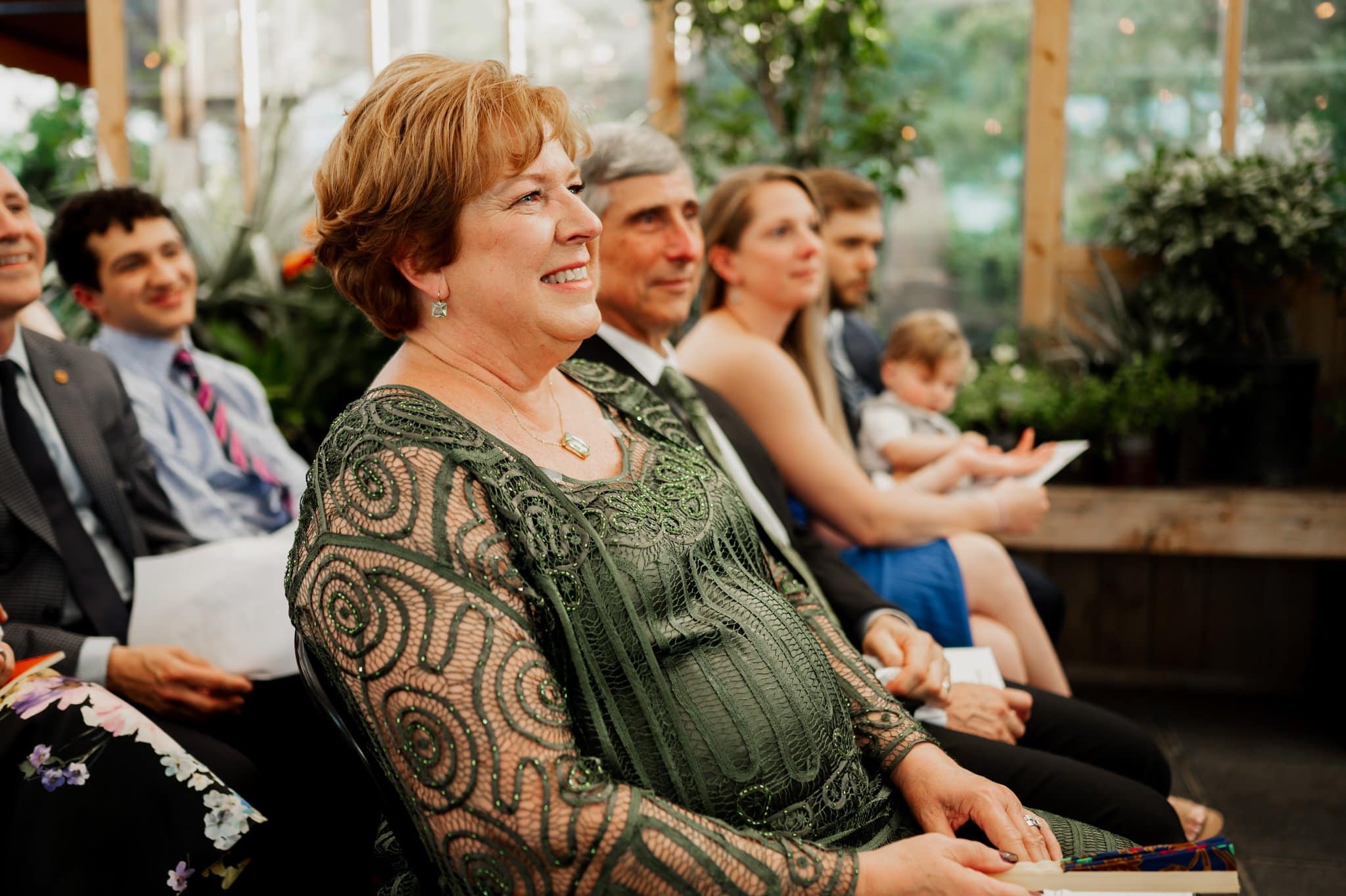 A woman in a green dress smiles while sitting among a group of people at an indoor wedding event, possibly featuring photos taken at Blockhouse.