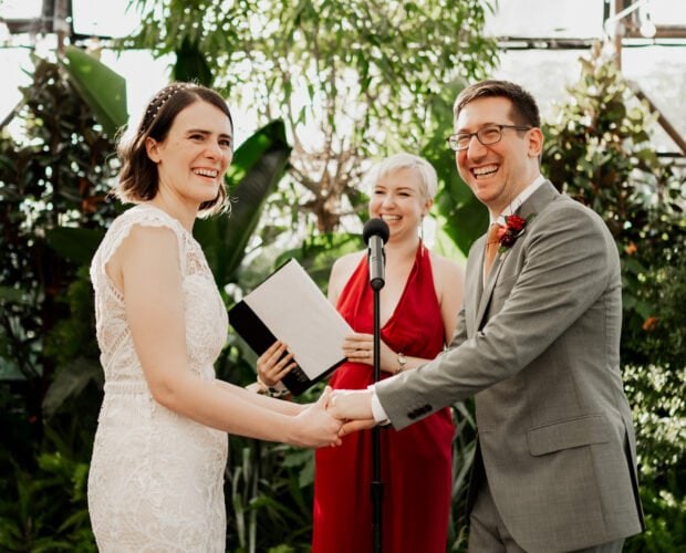 A couple holding hands and smiling during a wedding ceremony at Blockhouse, surrounded by a lush, green environment.